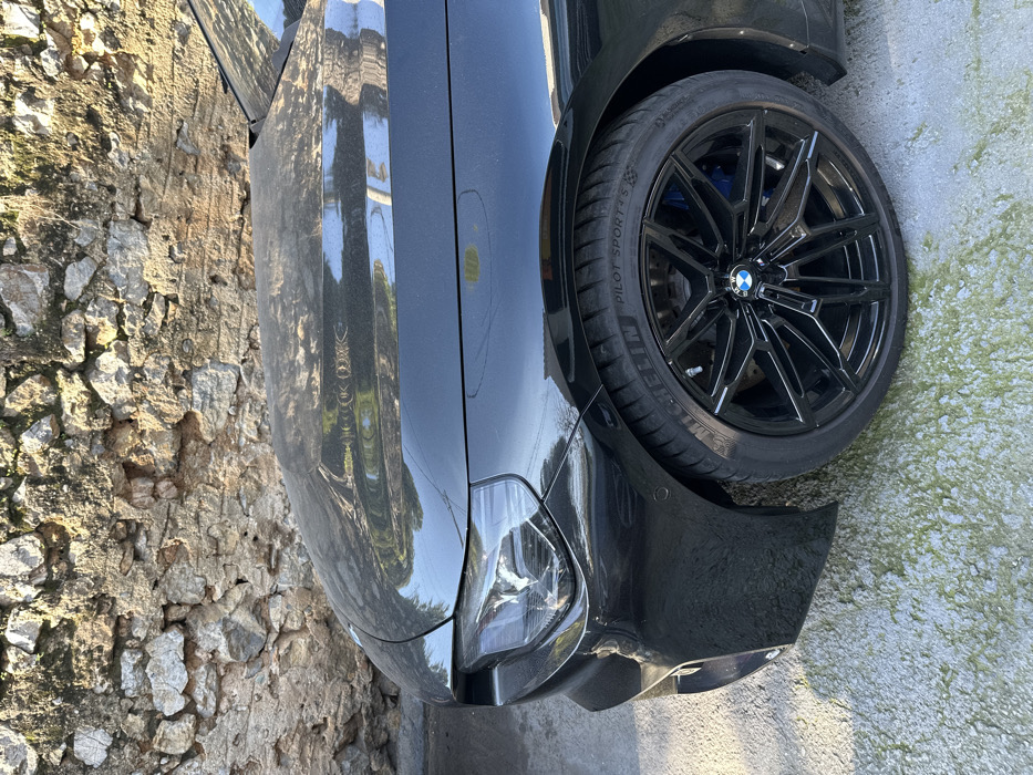 The image captures the front quarter of a black BMW car, parked outdoors. The car's glossy black paint reflects the bright surroundings, including hints of trees and sky. It is equipped with black multi-spoke alloy wheels, featuring a silver BMW logo in the center cap, and blue brake calipers are visible through the spokes. The tires are branded "MICHELIN PILOT SPORT 5".
To the left of the vehicle, there is a rugged, light-colored stone wall, constructed from uneven, stacked stones. The ground directly beneath the car is a light-toned, possibly paved surface, with patches of green moss or algae visible, especially near the wheel. The scene is illuminated by bright sunlight, indicating daytime conditions. The car appears stationary, with no activity taking place. The setting, characterized by the stone wall and outdoor light, suggests a location in Metamorfosi, Greece.