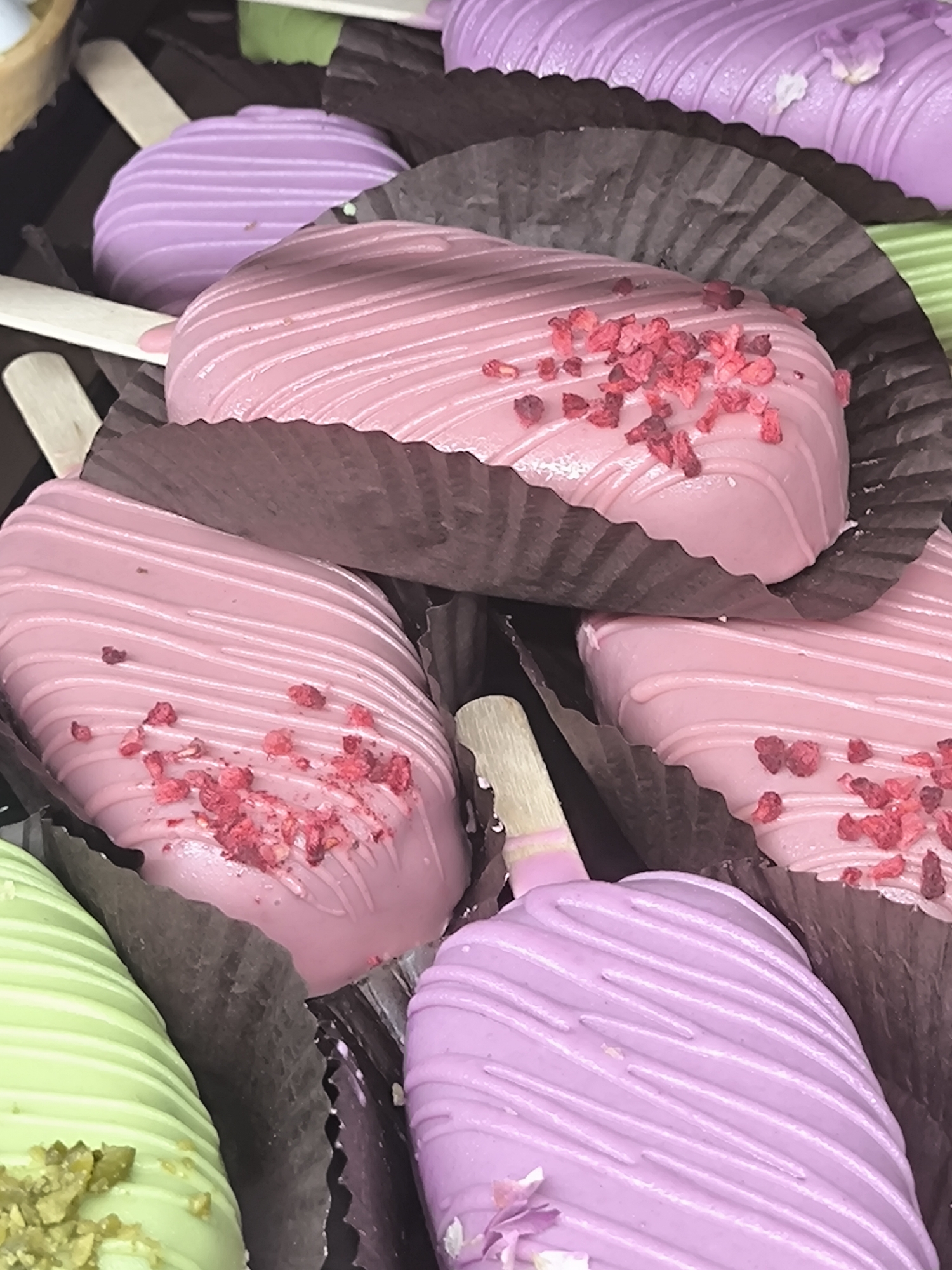 The image displays a collection of ice cream pops arranged in what appears to be a display case. The pops are various pastel colors like pink, purple, and green, each coated with a smooth, glossy exterior that has thin, wave-like lines running across the surface. Some of the pink pops are sprinkled with small, red, granular toppings. They are held in individual dark brown paper cups or liners with a rippled edge, and each has a wooden stick inserted. The pops are closely packed, obscuring the full view of each. The photo exhibits a close-up perspective, focusing on the texture and color details of the desserts. There is no direct indication of Riga, Latvia, in the image.
