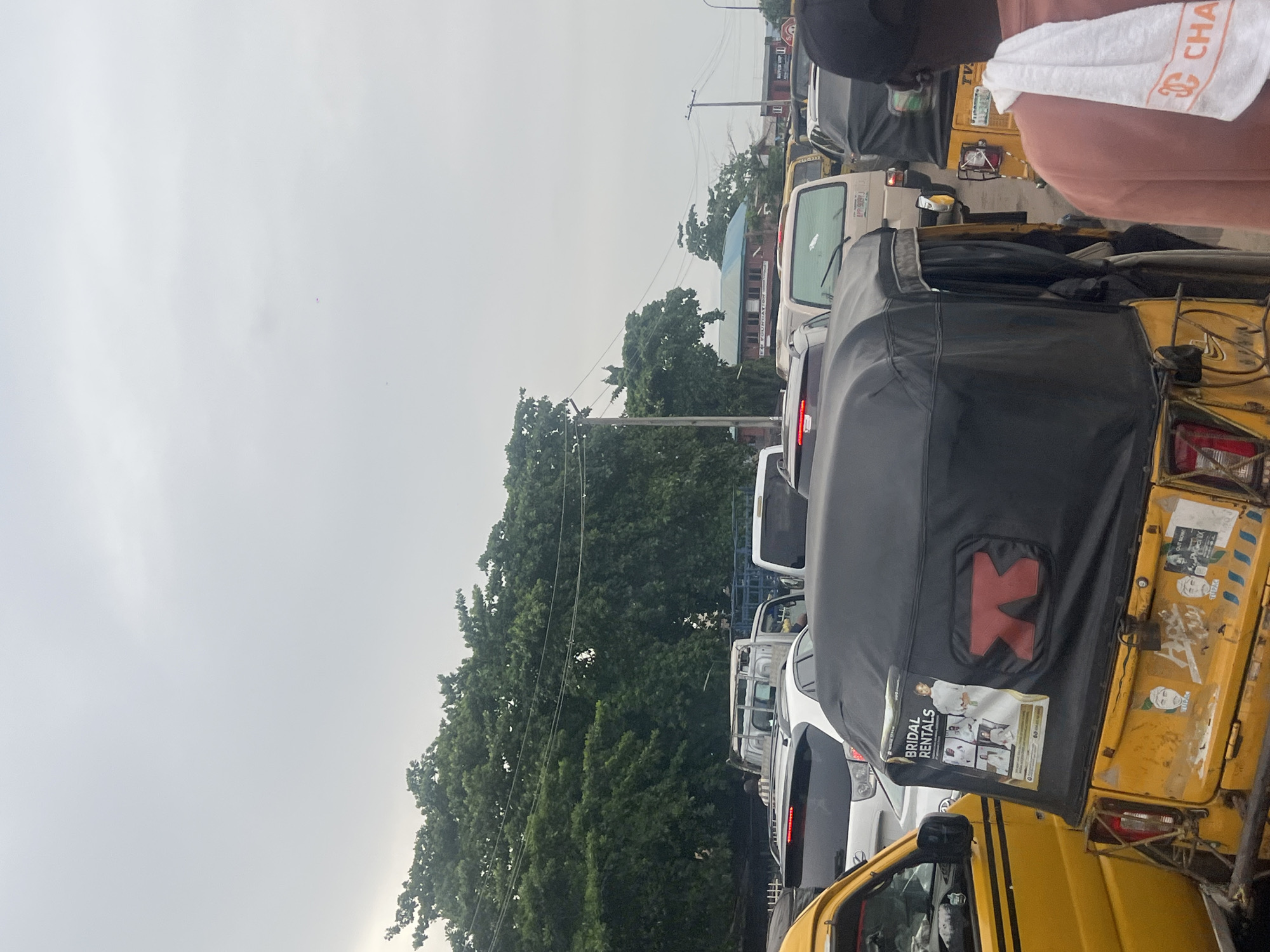 The image depicts a street in Alasia, Nigeria, congested with vehicular traffic. Predominantly yellow 'keke' (auto-rickshaws) are interspersed among other cars, including white sedans and minivans. The vehicles are in a standstill, suggesting a traffic jam. Lush green trees flank the road, partially obscuring buildings in the background. Utility poles with wires are visible. The sky is overcast. A person is partially visible in the foreground with a branded towel over the shoulder. Advertisements are affixed to the rear of one of the 'keke'.