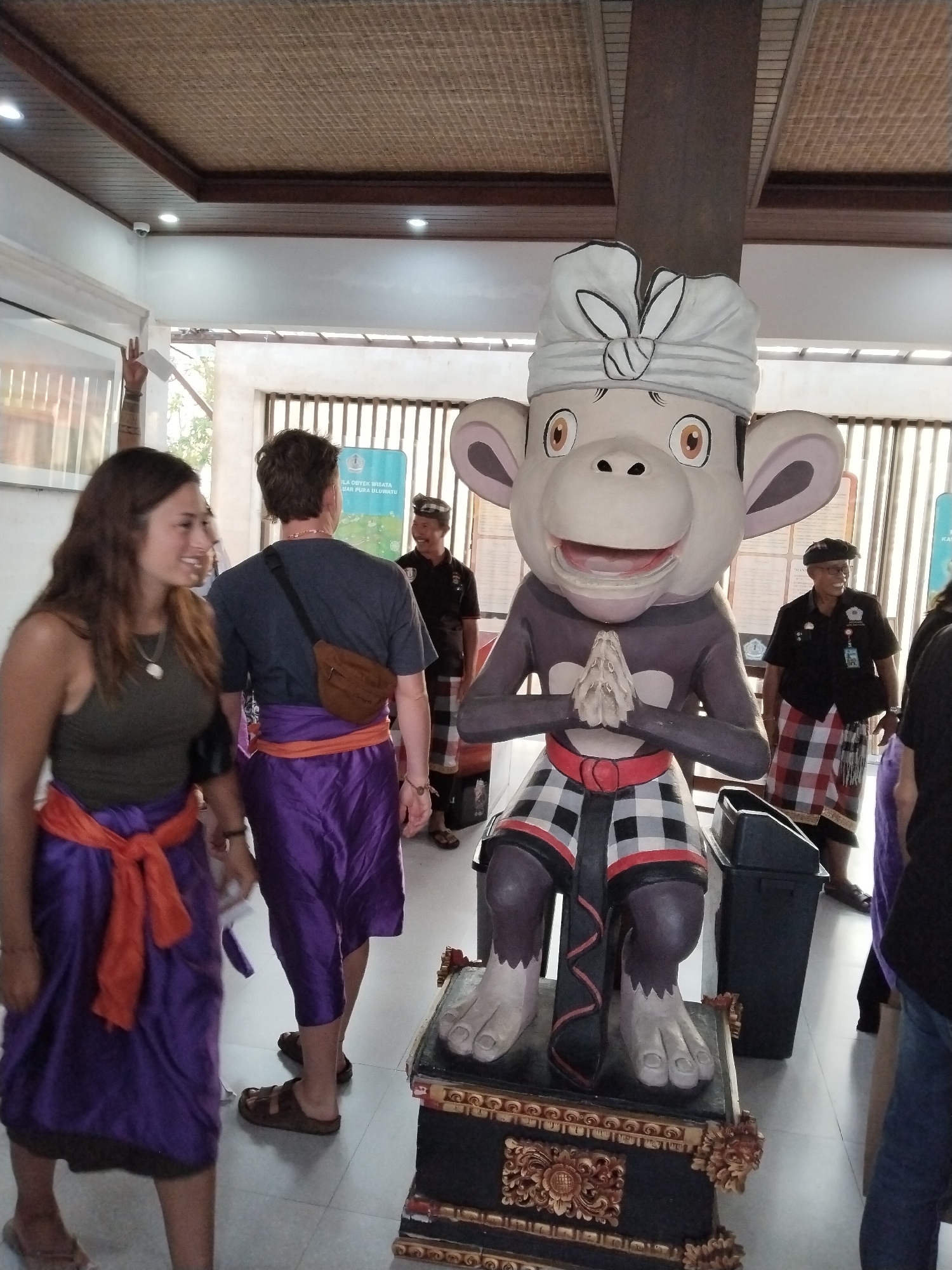 In Kuta Selatan, Indonesia, a large, cartoonish monkey statue with a white headwrap and clasped hands stands prominently in an indoor space with a thatched ceiling. Two women in purple sarongs and orange sashes are in the foreground, facing a man in a dark t-shirt with a brown fanny pack and a purple sarong. In the background, two men wearing traditional checkered sarongs and dark shirts are visible, one of whom is smiling. A black trash can is positioned near the monkey statue, and the environment appears to be a public indoor area, possibly a tourist attraction or cultural center.