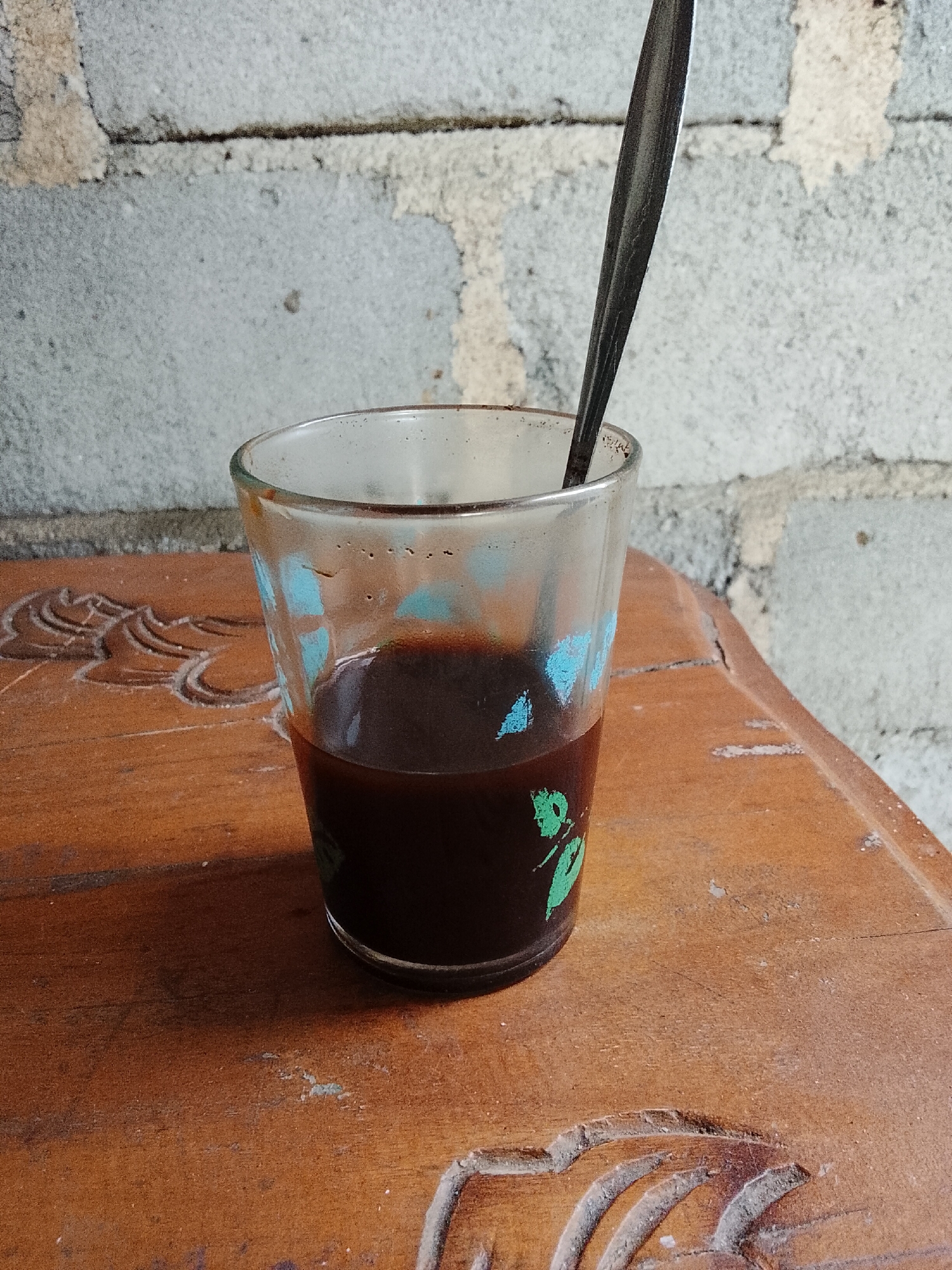 A close-up, eye-level shot shows a glass of dark liquid, likely coffee, sitting on a wooden surface. A metal spoon is partially submerged in the liquid, its handle extending out of the glass. The glass has a faint blue and green design around its base. The wooden surface appears worn and features carved patterns. In the background, a textured wall made of light-colored bricks is visible. The lighting suggests an outdoor or well-lit indoor environment. The scene is static and does not depict any activity or individuals. The location is identified as Padang Dalom, Indonesia.