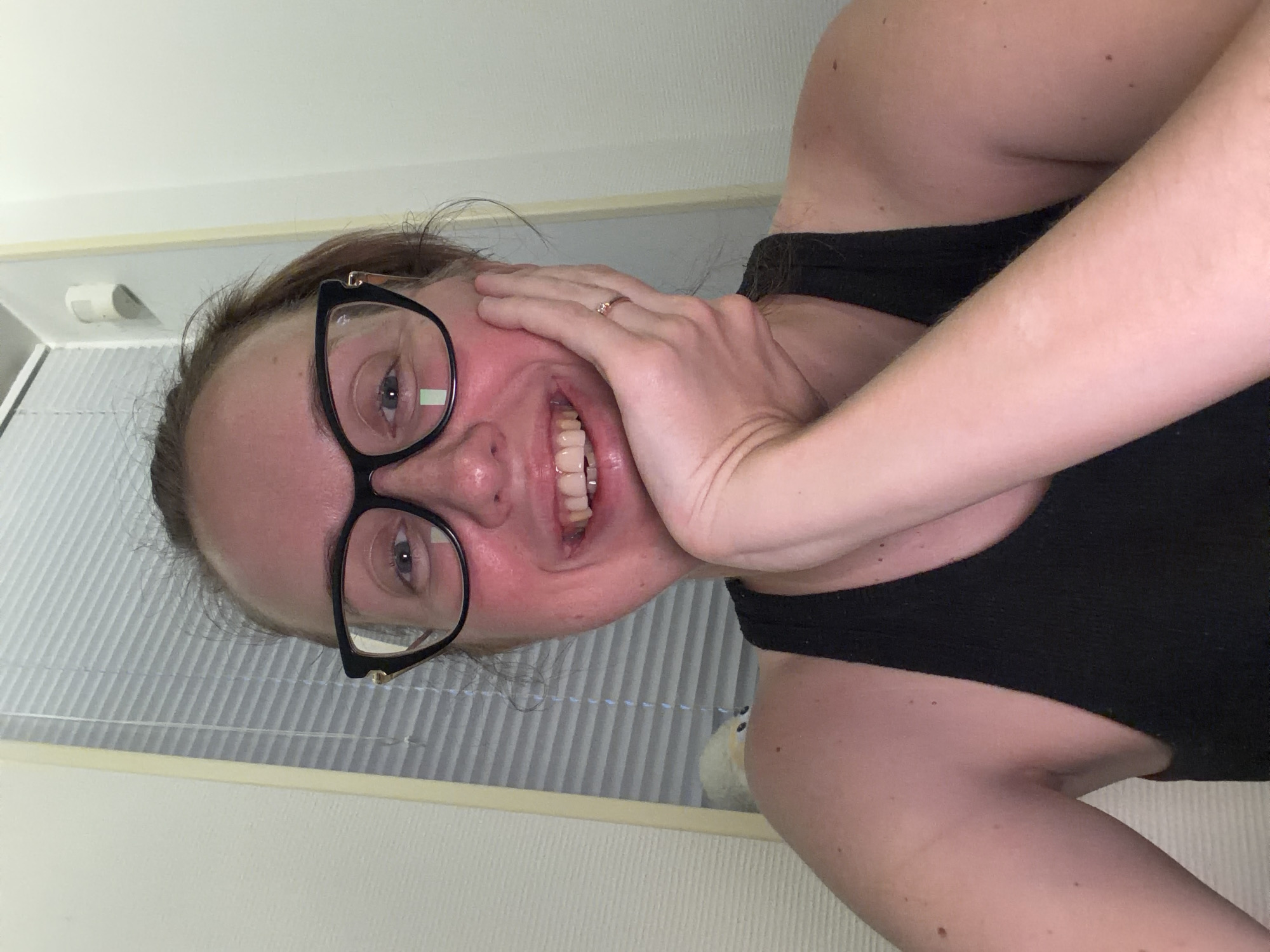 A woman with light skin and auburn hair pulled back is looking at the camera and smiling. She is wearing black-rimmed glasses and a black tank top. Her right hand is resting on her chin, and she is wearing a ring on her ring finger. The background is a wall with white blinds and a white light fixture.