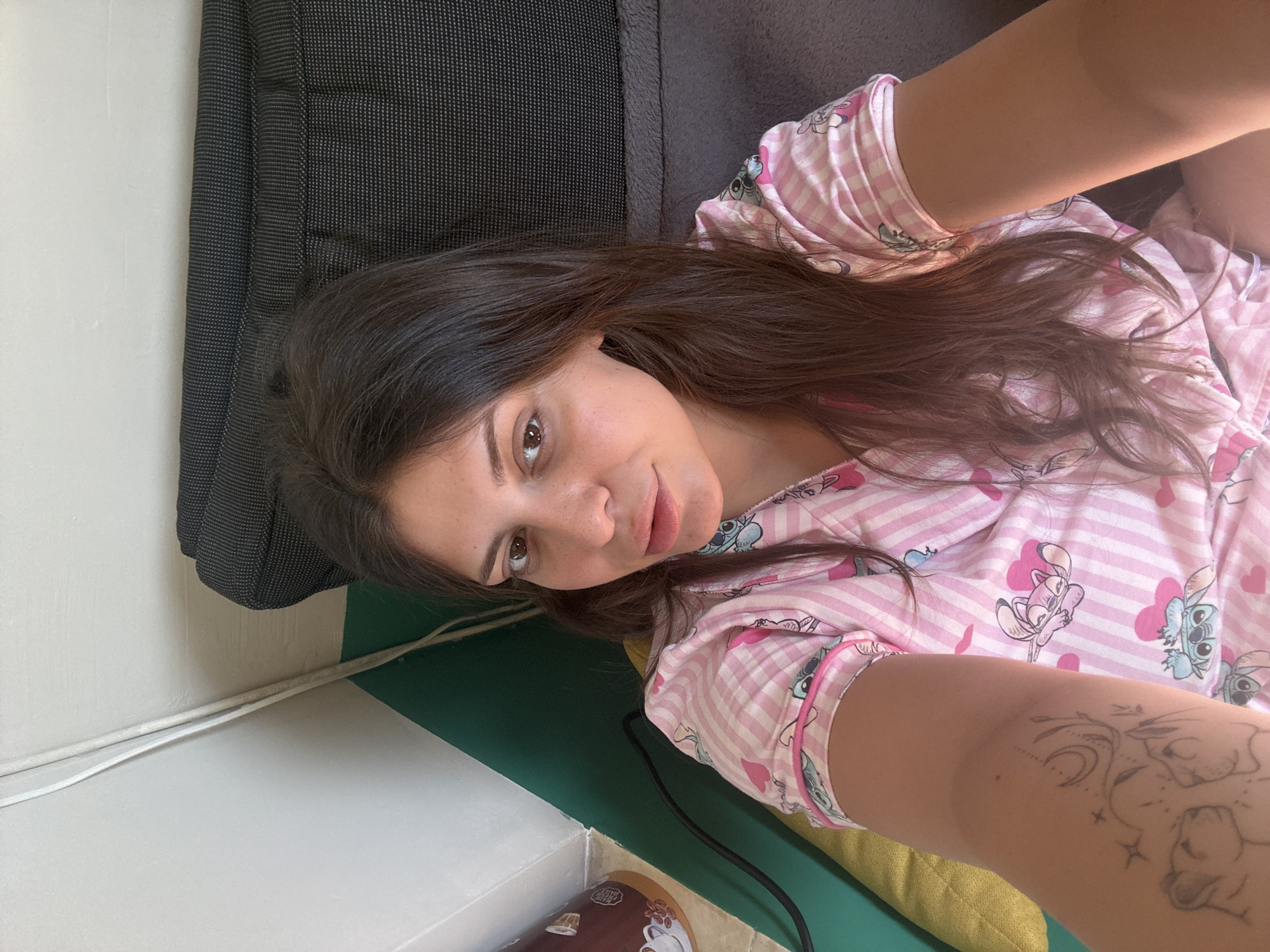 A young woman with long, dark hair and brown eyes is lying down and taking a selfie. She is wearing pink and white striped pajamas with images of Stitch from Lilo & Stitch. Her left arm, which is visible in the foreground, has a tattoo of celestial elements and two lions. The background is out of focus, but appears to be an indoor setting with a textured dark gray surface, a green cushion, and a white wall with a white cord running along it. The scene does not contain any discernible Plovdiv, Bulgaria landmarks or elements.