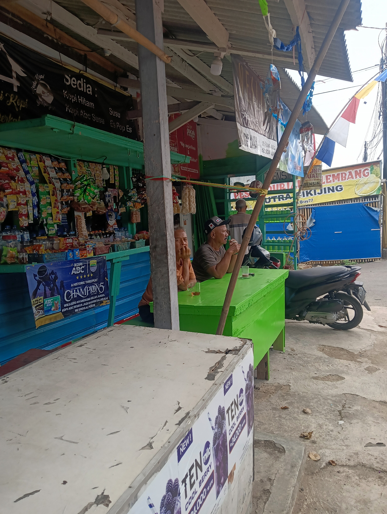 A street-side kiosk is shown in Manggari, Indonesia, selling various goods. The kiosk has a green structure with shelves stocked with snacks and drinks. A man in a brown shirt sits at a green table in front of the kiosk, looking away from the camera. Another man, wearing a black cap and brown shirt, is also seated at the table, facing left and appearing to converse. A motorcycle is parked to the right of the table. Several banners and flags are displayed, including one with an image of a durian fruit and text indicating fuel sales. Another banner in front of the kiosk advertises "Kopi ABC Champion 5". The ground is paved with concrete, and there are some debris and leaves present. The overall scene depicts a typical roadside establishment.
