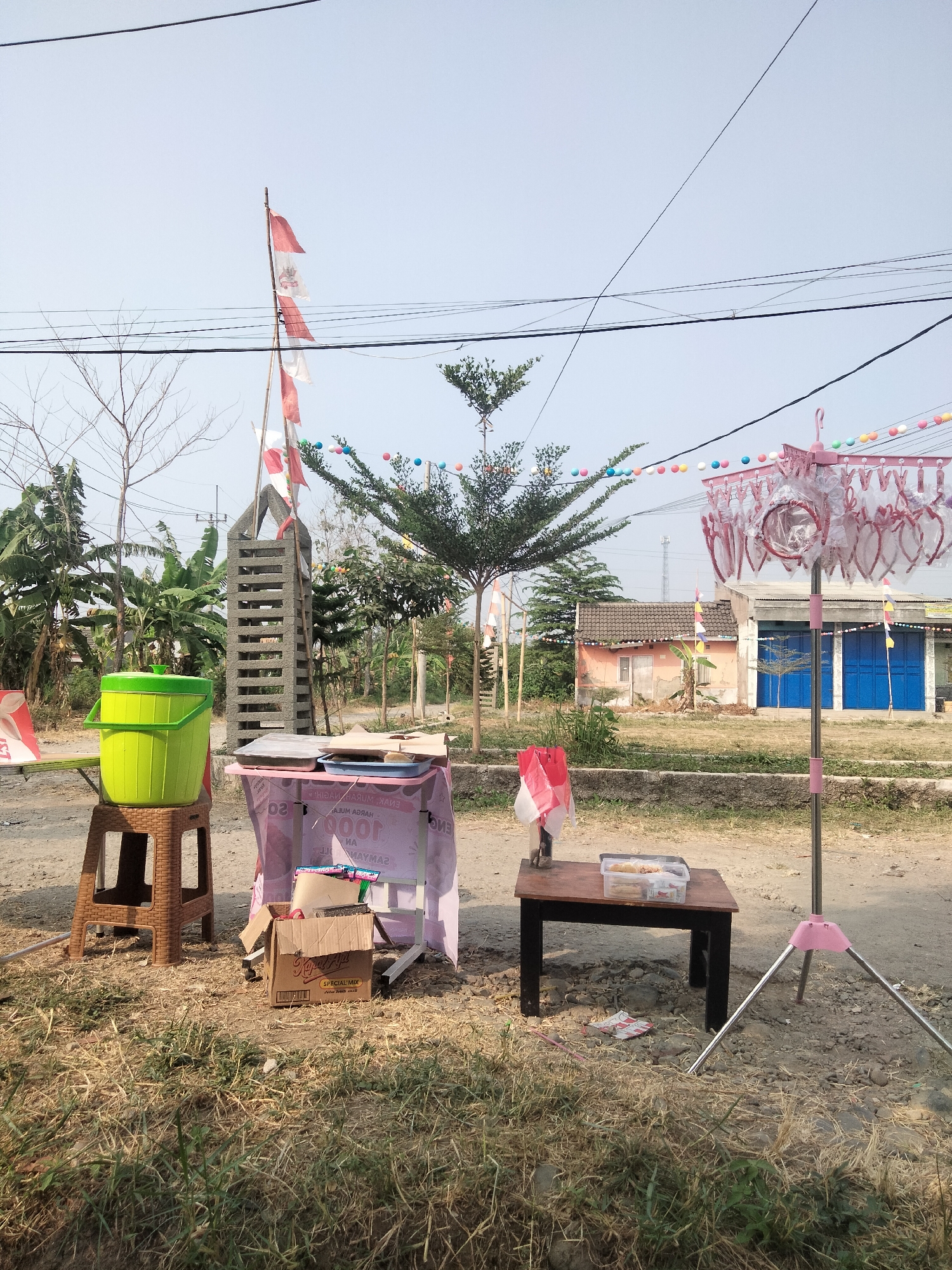 The image depicts an outdoor scene in the city of Cariu, Indonesia, featuring a temporary roadside stall. In the foreground, a brown plastic stool supports a bright green bucket with a lid. Next to it, a pink tablecloth covers a table, on which various items are placed, including what appears to be food wrapped in paper and a pink tray. A cardboard box is situated on the ground in front of this table. To the right, a smaller dark wooden table holds a clear plastic container with food items, possibly snacks. A pink clothes drying rack with clothespins stands nearby, adorned with colorful string lights. A tall, textured concrete structure resembling a layered pillar stands behind the green bucket. Indonesian flags, red and white, are displayed on poles and attached to strings of colorful decorative lights that stretch across the scene, including some hanging from overhead wires. In the background, a modest building with a pinkish-orange facade and a dark roof is visible, alongside a building with blue double doors. The ground is a mix of dirt, gravel, and dry grass. The overall impression is that of a small, informal vending setup, possibly for a local event or market.