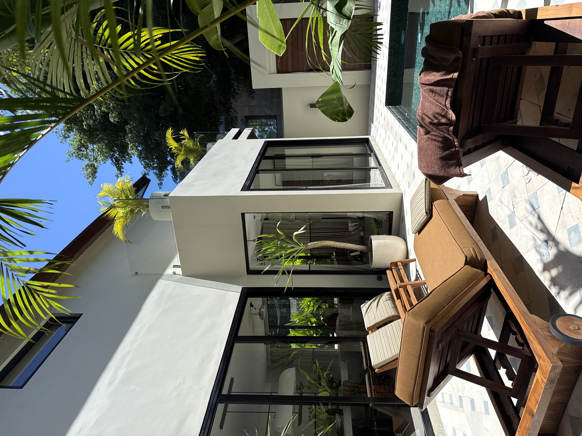 The image captures a bright, sun-drenched outdoor scene of a modern architectural building and an adjacent patio with a swimming pool, characteristic of a tropical resort or villa in a location like Kuta Selatan, Indonesia. The perspective suggests a clear, sunny day, with strong natural light casting distinct shadows.
In the foreground and along the left, vibrant green palm fronds and other large tropical leaves frame the view, creating a lush, natural canopy. The central focus is a contemporary building featuring clean, white or light-grey stucco walls contrasted with prominent black-framed windows and glass doors. These reflective surfaces mirror the surrounding greenery and clear blue sky.
An inviting outdoor area unfolds on the right, paved with light-colored tiles that feature a subtle geometric pattern, possibly in shades of white and light blue. This patio is furnished with several pieces of wooden outdoor furniture, including lounge chairs with both striped and solid brown cushions, and a low wooden side table or cart. A brown towel is draped over one of the lounge chairs, hinting at recent or imminent use.
Adjacent to the tiled patio is a swimming pool with clear, turquoise-blue water, reflecting the daylight. In the background, beyond the pool, another section of the building displays dark wooden paneling, adding a contrasting texture. A large potted plant, resembling a Dracaena, with a long stem and green leaves, stands near the building's windows.
The scene conveys a sense of tranquility, luxury, and tropical relaxation. No individuals or visible text are present in the image.