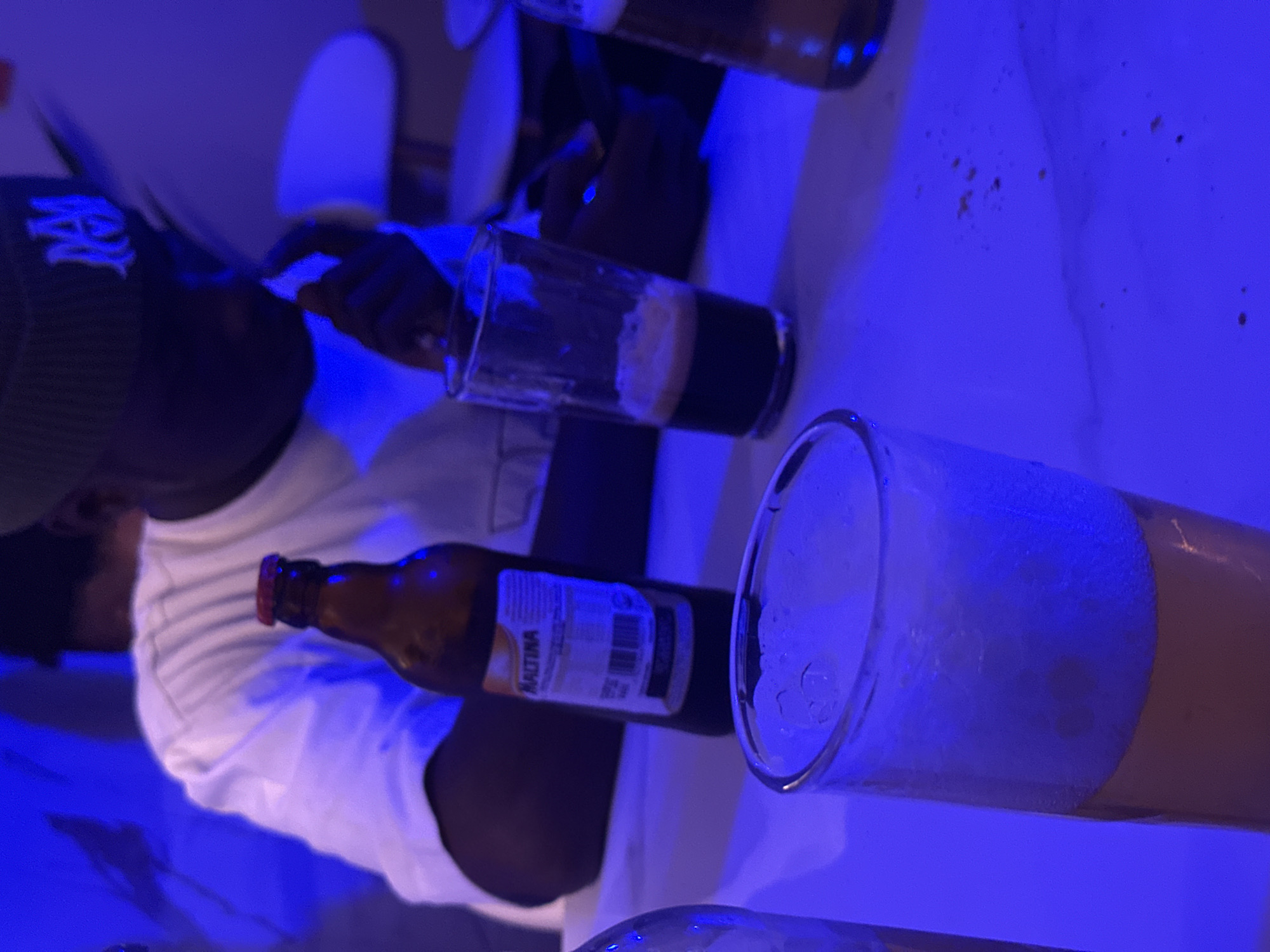 The image shows an individual wearing a dark-colored beanie with a white logo on the left side, a white t-shirt, and dark-colored skin. They are seated at a table with a white surface. On the table are several glasses containing liquids, likely beverages, and at least one bottle labeled "Maltina". The location is presumed to be Aba, Nigeria, based on the Maltina brand presence, though no other details definitively confirm this. One of the glasses contains a dark liquid with a lighter layer on top, possibly foam or froth. Another glass is filled with a light-colored beverage and a thick layer of foam. The lighting appears to be predominantly blue. The individual seems to be holding a glass. There appear to be chairs in the background.