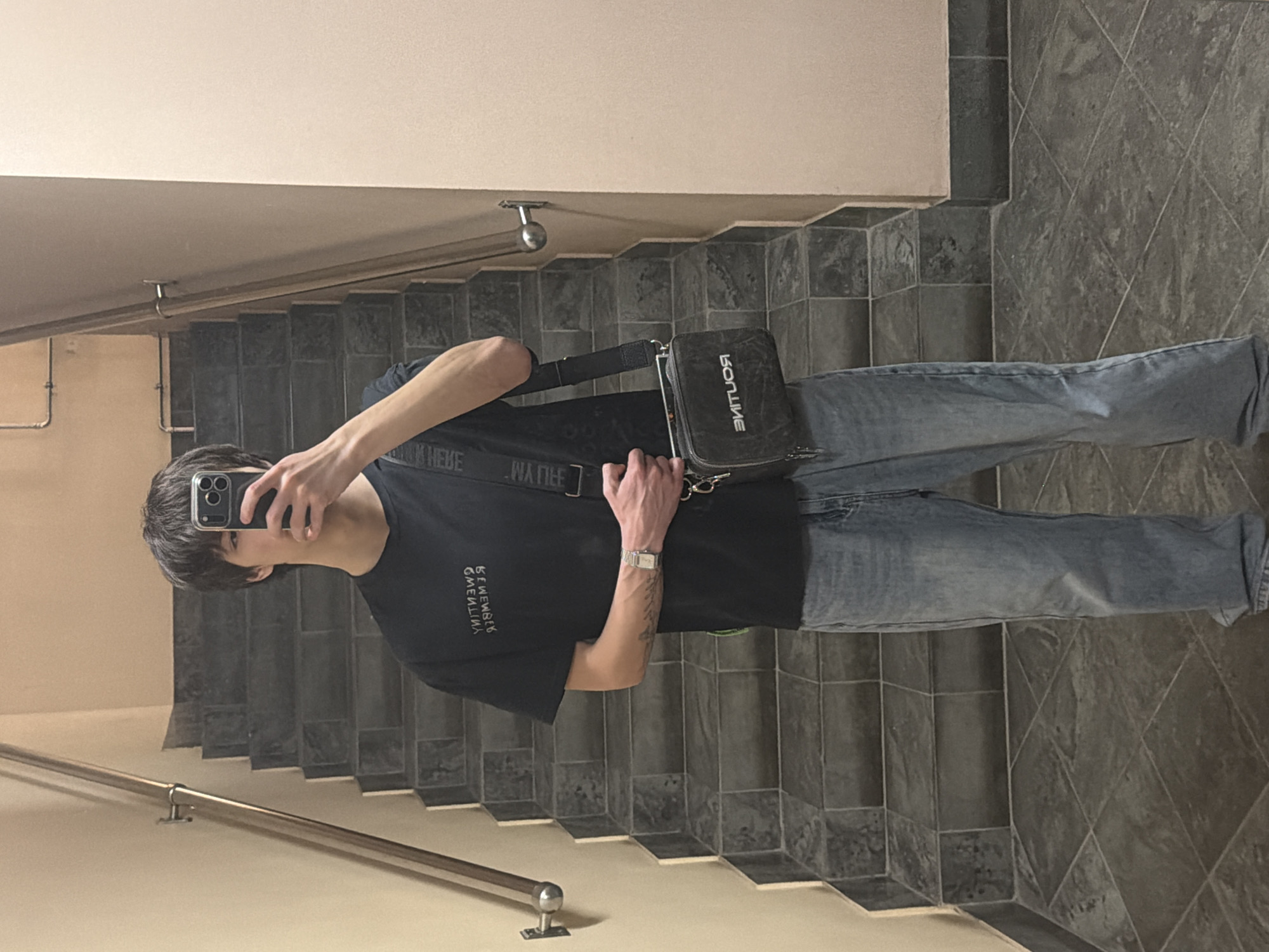 A young man is taking a selfie in a mirror on a staircase. He is wearing a black t-shirt with white lettering, light blue jeans, and a silver wristwatch. A black cross-body bag is slung across his torso. The staircase has grey tile flooring and a light beige wall. The handrail is silver. The setting appears to be an indoor public space. The location cannot be determined to be Yekaterinburg, Russia, from this image alone.