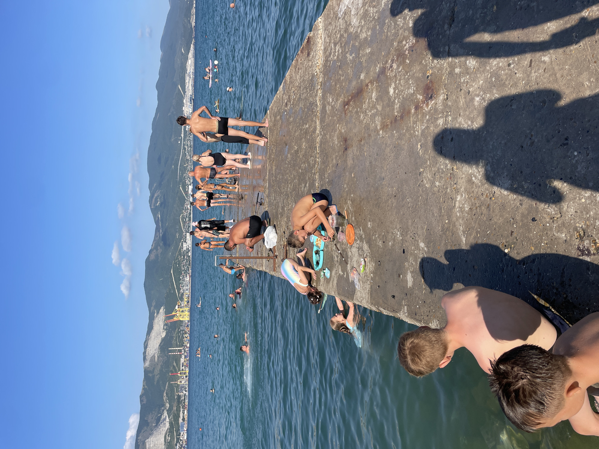 This is a photograph of a concrete pier extending into the sea. Several people are on the pier and in the water. In the foreground, two young boys are sitting on the edge of the pier, one looking out at the water and the other looking away from the camera. Behind them, a man is squatting down and looking at something on the pier. A girl in a light blue swimsuit is in the water to the left of the pier, and another girl in a multicolored swimsuit is leaning over the edge of the pier, also looking at something in the water. Further down the pier, a group of adults and children are standing and interacting. In the background, mountains and industrial structures, likely part of the city of Sadovyi, Russia, are visible along the coastline. The sky is blue with a few scattered clouds. The sea is calm and blue. The scene depicts a warm, sunny day at the beach.