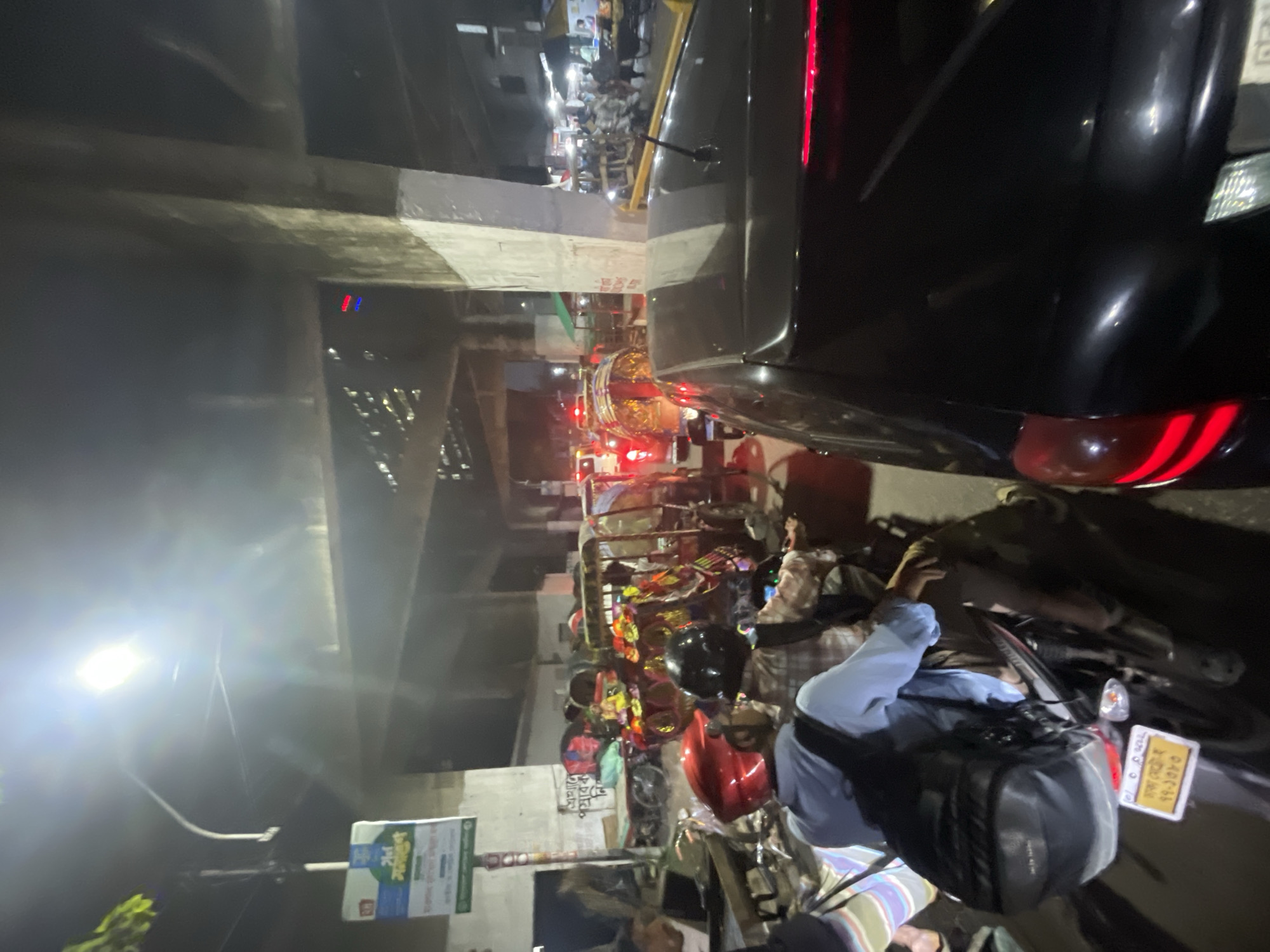The image shows a crowded street scene at night, likely in Dhaka, Bangladesh, based on visual cues and the style of signage. An elevated concrete structure, possibly a flyover or overpass, dominates the top-left of the frame. Heavy traffic, including cars and numerous motorcycles, is visible. A large black car is prominent on the right side, with its tail lights illuminated. A motorcyclist wearing a blue shirt and a black backpack is positioned in the lower right, seemingly waiting in traffic. A nearby motorcycle displays a license plate written in Bengali. Decorations hanging from a rickshaw or cart on the side of the road. A street light shines brightly on the left side of the image, washing out some details. A roadside sign with Bengali text is also visible.
