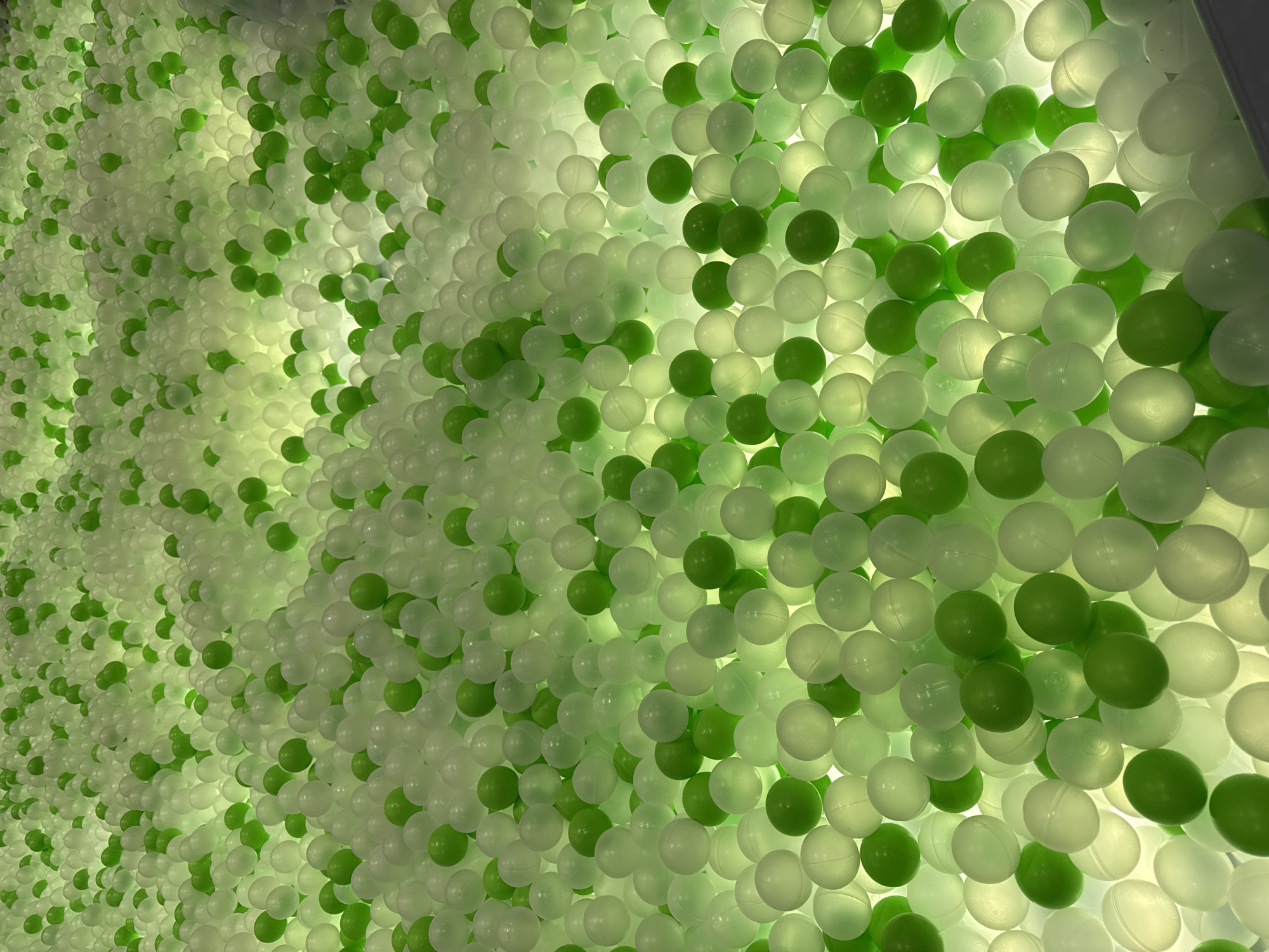 The image shows a dense wall filled with white and green plastic balls. The balls are packed tightly together and appear to be illuminated from behind, creating a glowing effect. No people are visible. The scene does not reveal any information that ties it to the location of Перовское сельское поселение, Ukraine.