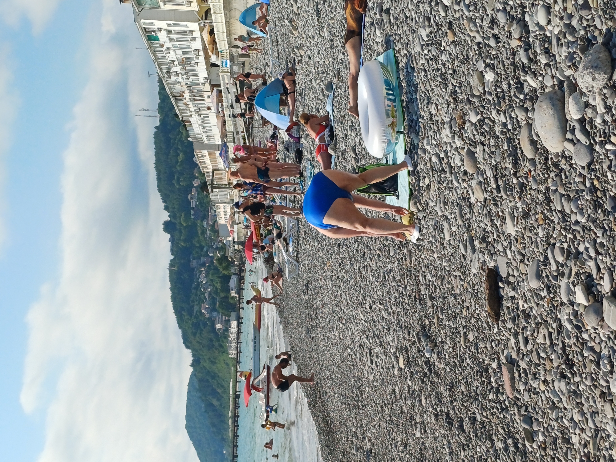 This media file captures a bustling scene at a pebble beach in Sochi, Russia, under what appears to be a bright, partly cloudy sky, suggesting a warm summer day. The image is oriented vertically, with the sea on the left and the beach and buildings on the right.
A diverse group of people are engaged in various beach activities. Some are sunbathing on towels and mats spread across the pebbles, while others are walking along the shoreline or wading into the shallow water. A prominent subject in the foreground is a person in a blue swimsuit performing a yoga pose. Elsewhere, people are relaxing under colorful umbrellas, and a few are seen entering or exiting the sea.
Objects visible include beach towels, umbrellas, and an inflatable ring. The background reveals a coastal town with multi-story buildings, characteristic of a resort area. A steep, tree-covered mountainside rises behind the buildings, a common geographical feature of the Sochi region. The presence of a pier or breakwater extends into the sea, with a few people on it.
The overall mood is one of leisure and recreation. The clear sky and numerous beachgoers suggest a peak season for tourism. No discernible text is visible in the image.