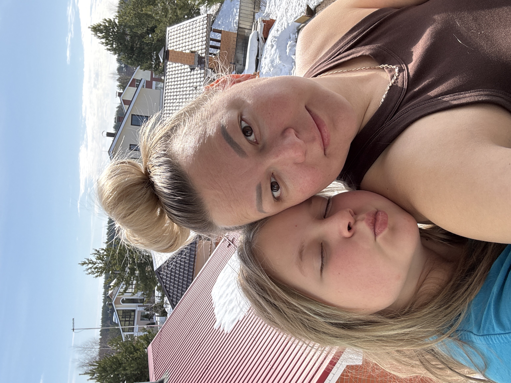 A daytime close-up selfie shows two individuals positioned against a residential and natural landscape in Русский Вожой, Russia. The primary subject is an adult female with blonde hair pulled into a high bun, wearing a dark brown tank top and a thin gold chain necklace. She looks directly at the camera with a neutral expression. Her left ear displays a small gold earring. To her right, a younger female, likely a child, with light brown hair and wearing a blue t-shirt, leans her head against the adult's shoulder. The child's eyes are closed, and her lips are puckered in a "kissy face" expression.
The background reveals a suburban environment from an elevated viewpoint. Immediately behind the subjects, a red corrugated metal roof with patches of snow is visible. Further back, multiple detached houses with varied architectural styles, including light-colored walls, dark window frames, and roofs made of brown tiles or red corrugated metal, are discernible. Patches of snow cover the ground and some rooftops, and numerous evergreen trees (pines, spruces) are interspersed throughout the scene, along with some bare deciduous trees. A tall, thin antenna-like structure is visible extending above one of the houses, and a brick chimney protrudes from another roof. The sky is bright blue with scattered white clouds, indicating clear weather and ample sunlight.