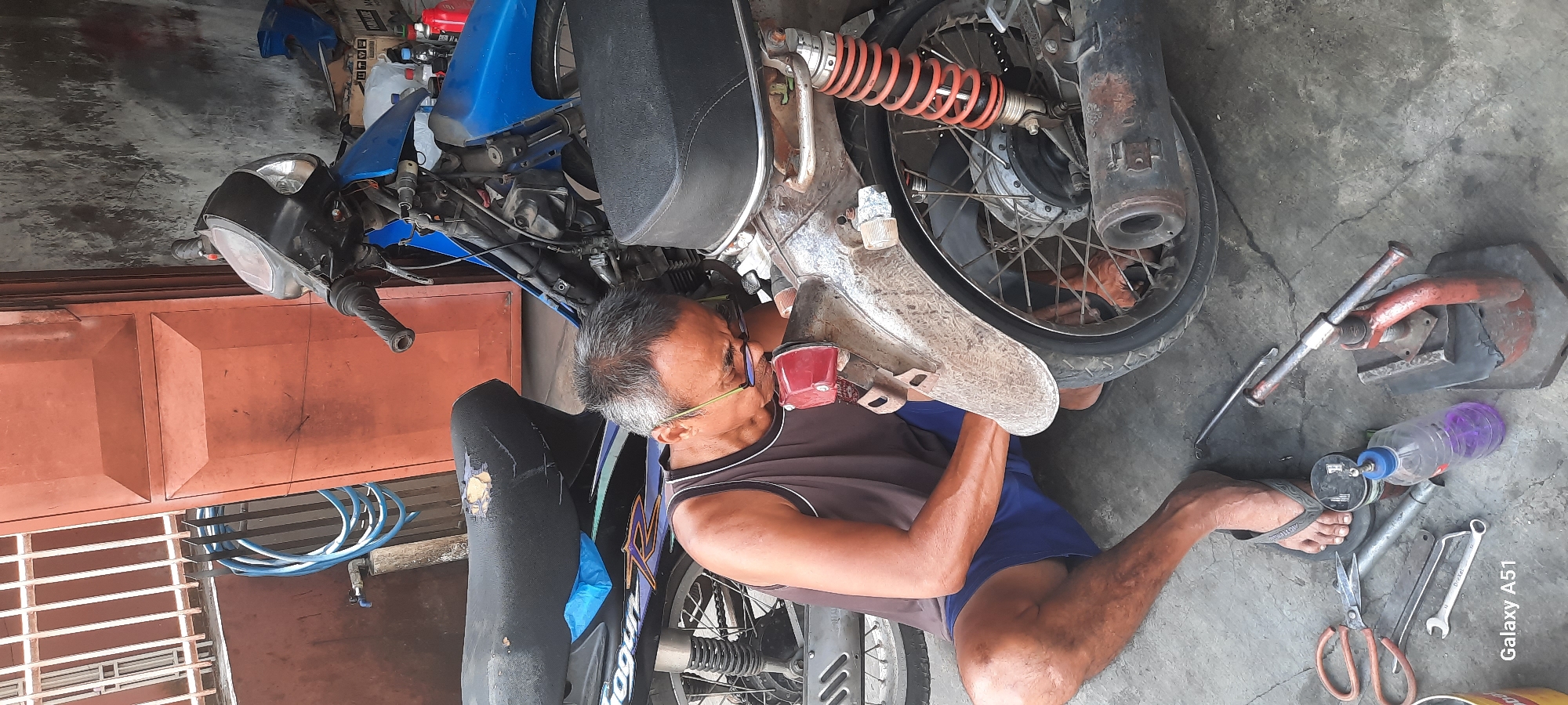 A middle-aged man with graying hair and glasses is sitting on the ground and repairing a blue motorcycle. He is wearing a brown tank top and blue shorts. The motorcycle is parked in front of a large orange garage door. Tools, including wrenches, pliers, and a bottle of purple liquid, are scattered on the ground around the man. The scene appears to be an outdoor workshop or garage in the city of Selopuro, Indonesia. The overall impression is one of a typical day in a local motorcycle repair shop.