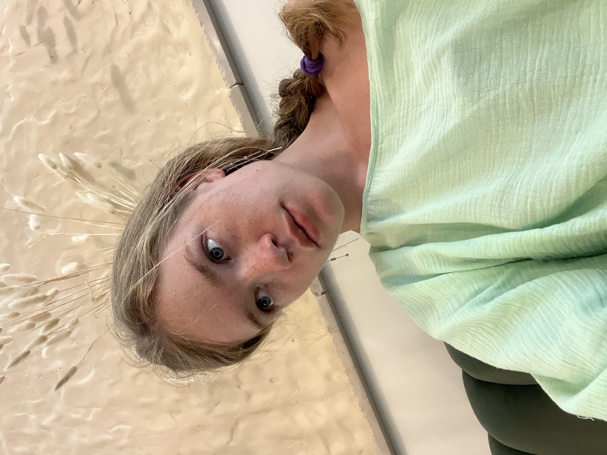 A young woman with blonde hair in a braid is sitting indoors. She is wearing a light green, textured blouse and is looking away from the camera. The background consists of a textured beige wall with a light fixture or decorative element made of dried grasses. There is also a glimpse of a dark green upholstered chair in the foreground. The image was taken at an eye-level perspective, with the subject occupying the majority of the frame. The lighting appears to be natural and soft.