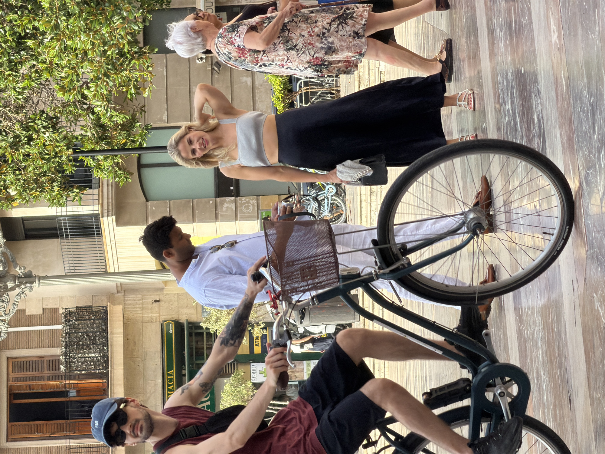 In the city of Unknown, Unknown, on a bright day, a man wearing a dark red sleeveless shirt, dark shorts, a blue baseball cap, and sunglasses is riding a bicycle. He has tattoos on his arms. The bicycle has a brown basket attached to the handlebars. Behind him, a man in a white shirt and white pants walks away from the camera. To the right of the cyclist, a woman with blonde hair is wearing a grey crop top and a long dark skirt, with a small black purse in her hand. Further to the right, an older woman with white hair is wearing a floral print dress and walking away. The background shows a building with windows and balconies, and trees with green leaves. The ground is paved with a reflective, marble-like surface.