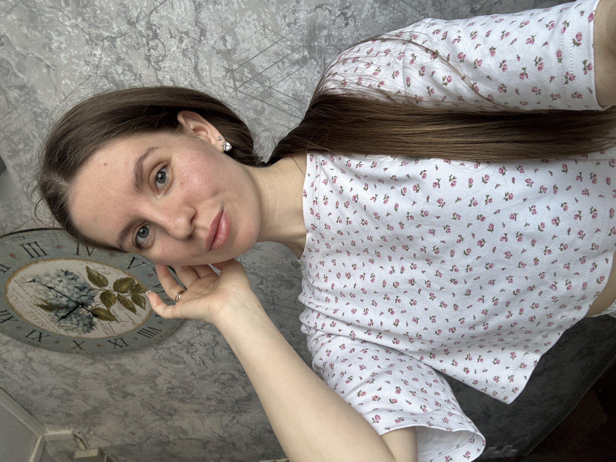 A woman is depicted in a selfie, presumably indoors. She has long, straight brown hair, fair skin, and light-colored eyes. She is wearing a white top patterned with small pink flowers and a small earring. Her left hand is near her face, and she is wearing a ring on her finger.
In the background is a wall with a gray, textured pattern, and an ornate clock with Roman numerals. The clock features a floral design in the center.
The image may have been taken in Klin, Russia, but the provided image does not contain information to confirm this.