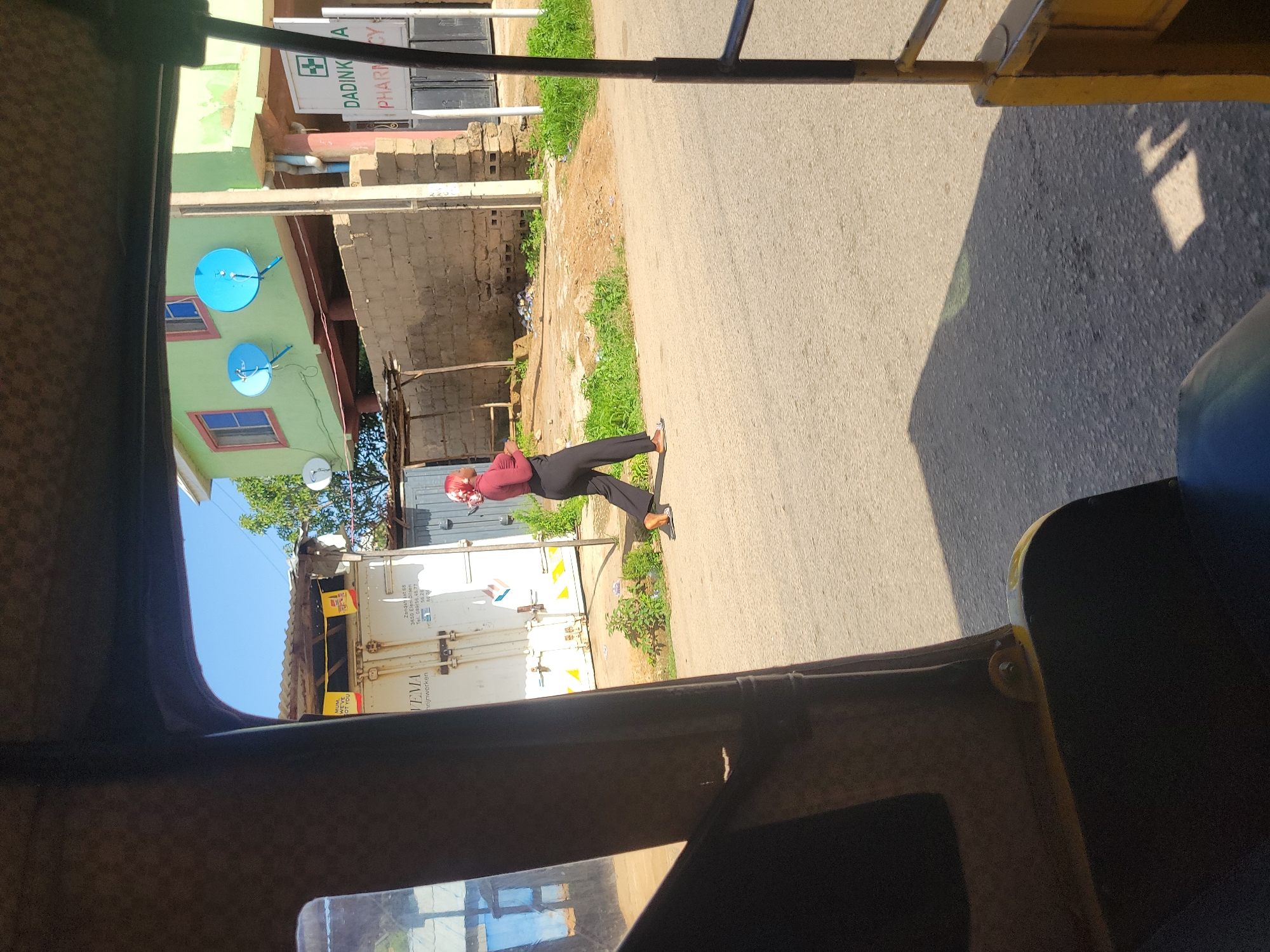 The image shows a street scene in Jos, Nigeria, viewed from inside a yellow auto-rickshaw. A woman wearing a maroon hijab and a maroon top with black pants is walking on the side of the road. In the background, there is a light green building with two satellite dishes mounted on its wall. Adjacent to the building is a sign for "DADINKA PHARMACY." To the left of the building, a white shipping container is visible, with the word "VEMA" printed on it. The street is paved, and there is green vegetation along the side of the road. The sky is clear and blue.