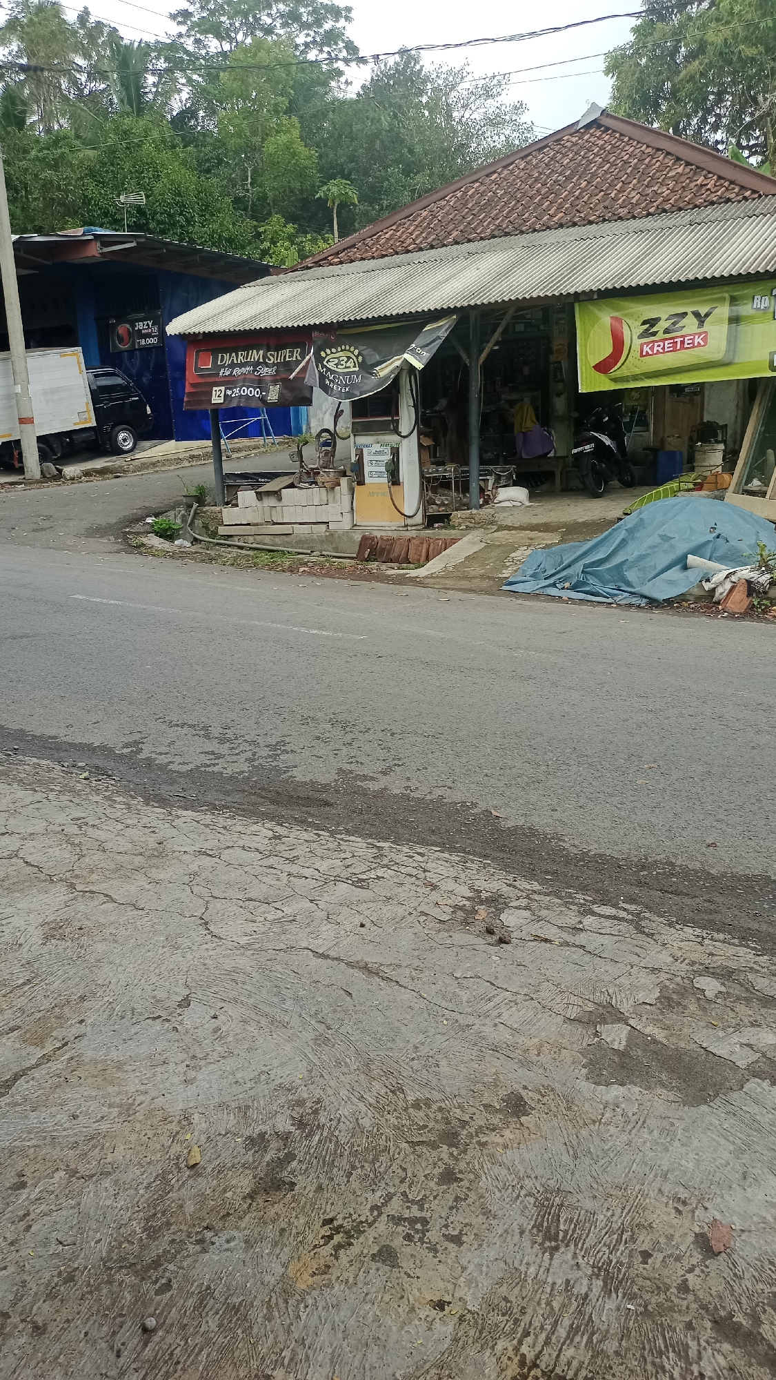 The image captures a roadside scene in Karyamandala, Indonesia, featuring a small roadside structure that appears to be a shop or warung. In the foreground, a paved road with visible cracks and patches of wear is prominent. To the right of the road, a blue tarp covers a pile of debris. Behind this, a building with a corrugated metal roof extends to the right.
The focal point of the scene is the roadside structure. A white and yellow gasoline pump is situated next to a collection of stacked white bricks and some brown, cylindrical objects, possibly fuel containers. Above the pump, a gray corrugated metal awning extends outwards, providing shade. Mounted on poles supporting the awning are several signs. One prominently displays "DJARUM SUPER" in red and white lettering, with "MAGNUM KRETEK" below it. Another sign, a bright green banner with a red J-shaped logo and white text reading "ZZY KRETEK," is visible on the right side of the structure.
To the left of the gasoline pump, a blue building is visible, with a white delivery truck parked in front of it. The blue building has signage, including one that appears to advertise cigarettes, with text such as "JAZY RAK 12" and "18.000". A black motorcycle is parked under the awning of the structure on the right. The background consists of lush green trees and foliage, suggesting a rural or semi-rural setting. The lighting indicates it is daytime, with overcast conditions. No people are directly visible in the image.