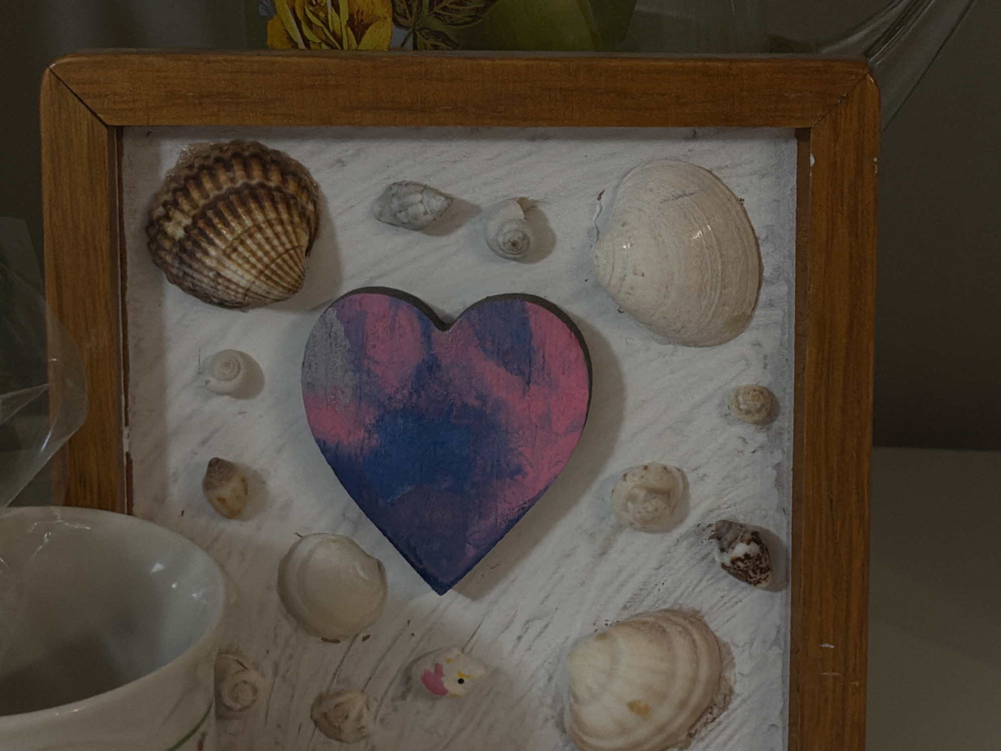 The image depicts a close-up view of a framed decorative display in an indoor setting. No specific location within the city of Sofia, Bulgaria, can be determined from the visual information provided.
The primary subject is a rectangular wooden frame, light brown in color with mitered corners, enclosing a light-colored, possibly textured, background. Mounted within this frame are approximately 15-20 seashells of various sizes, shapes, and natural hues, including ribbed brown and cream bivalve shells, smooth white bivalve shells, and smaller white and grey spiral shells. Centrally positioned within the frame is a heart-shaped wooden cutout, painted with a blended, mottled pattern of pink and blue. A small, white, bird-like figurine with a pink accent is affixed among the shells in the lower-central area of the frame.
In the bottom-left foreground, partially visible and out of focus, is the upper rim and interior of a white ceramic cup or bowl. The background behind the framed object is out of focus, showing indistinct elements including a yellow floral pattern on the top left and a green object towards the top right. No individuals, actions, or interactions are present in the scene.