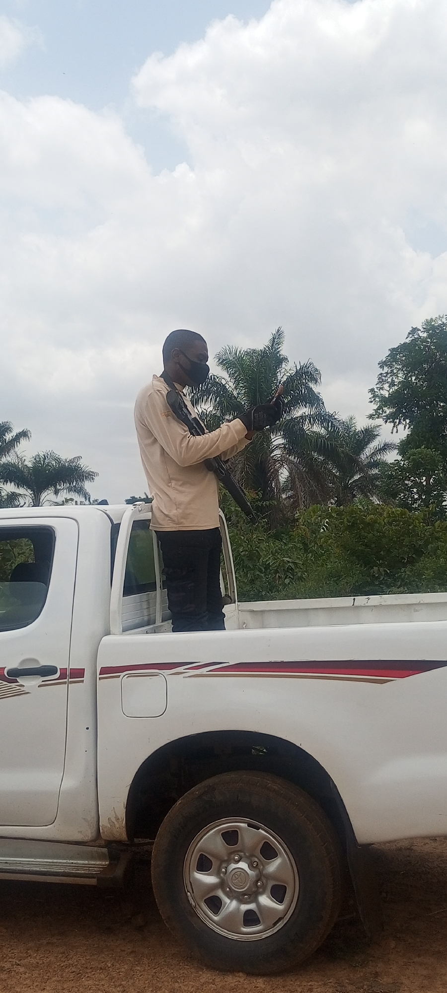 The image captures a daytime scene in what appears to be a rural or semi-rural area of Otunade, Nigeria. The central subject is a man standing in the bed of a white pickup truck, identifiable as a Toyota Hilux by the emblem on the wheel hubcap. He is wearing a light-colored long-sleeved shirt, dark trousers, and a black face mask covering his mouth and nose. The man is holding a dark long gun, resembling a rifle or shotgun, which is slung across his body. His gaze is directed downwards, suggesting he is looking at something held in his hands, possibly a mobile phone or another device.
The pickup truck is white with distinctive red and gold/brown striped decals running along its side. The ground beneath the vehicle appears unpaved, consistent with a dirt road or track. The background is dominated by lush green vegetation, including tall palm trees and dense foliage, indicating a tropical or subtropical environment. The sky above is bright and partly cloudy with large white cumulus clouds.
The activity depicted suggests a security presence, a patrol, or an escort, given the armed individual positioned in the back of the vehicle in an outdoor setting. The time of day is clearly daylight, likely mid-morning or early afternoon, with ample natural light and no harsh shadows. The man's expression is not visible due to the mask, but his posture appears calm and focused. No other individuals or specific events are clearly discernible in the frame.