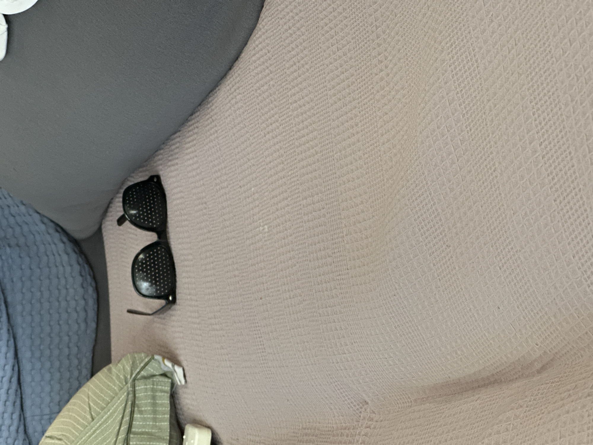 The image shows a pair of black pinhole glasses lying on a textured, light pink blanket. The glasses are positioned in the center-left of the frame. To the left of the glasses, a portion of a light green, striped pillow is visible. In the background, a dark gray pillow or cushion occupies the right side of the frame, with a blurred blue textured surface on the upper left. The scene appears to be indoors, possibly on a couch or bed. There are no people present in the image. The city of Nevinnomyssk, Russia, is not discernible from the visual information provided in the image.