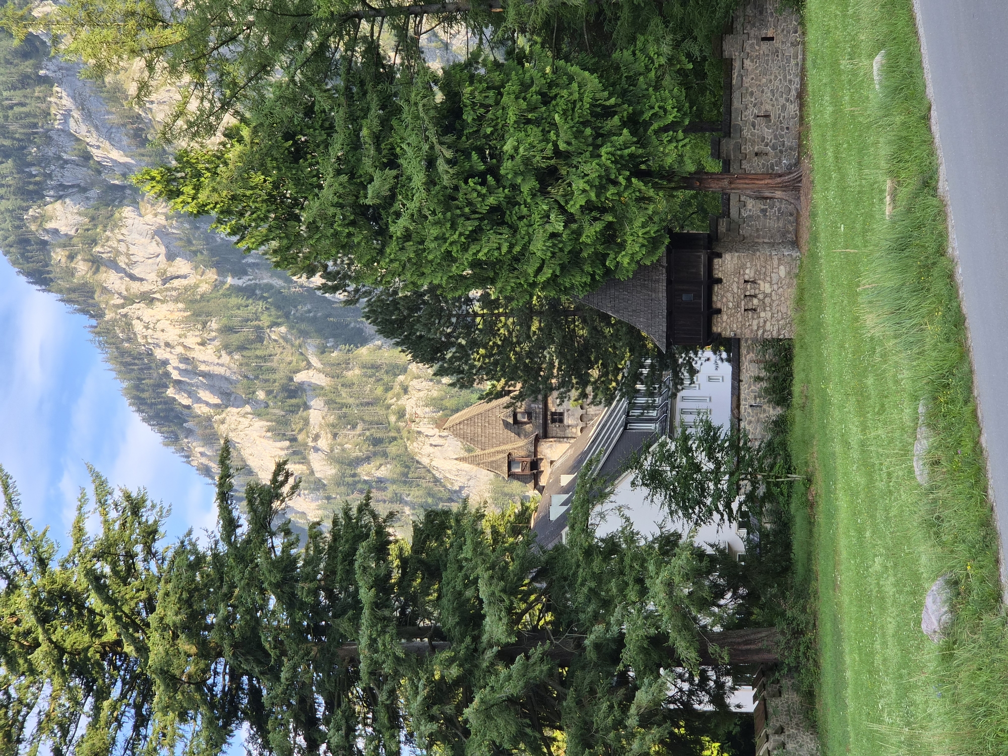 The image depicts a scenic view of a historic building and its surroundings, located in Eisenerz, Austria. In the foreground, a lush green lawn is dotted with rocks and bordered by a grey paved surface. Behind the lawn, a stone wall stretches across the scene, partially covered by vegetation. A small, dark wooden structure with a pointed roof and narrow windows stands atop the wall, appearing to be a watchtower or turret. Further back, a larger white building with a dark roof and multiple windows is visible, peeking through tall evergreen trees. These trees, along with other foliage, frame the scene, creating a sense of depth and enclosing the structures. In the background, a majestic mountain range rises steeply, its rocky face dotted with sparse trees and illuminated by sunlight. The sky above is a clear blue with a few wispy clouds. The overall impression is one of historical architecture nestled within a dramatic natural landscape, likely a part of the historic mining town of Eisenerz.