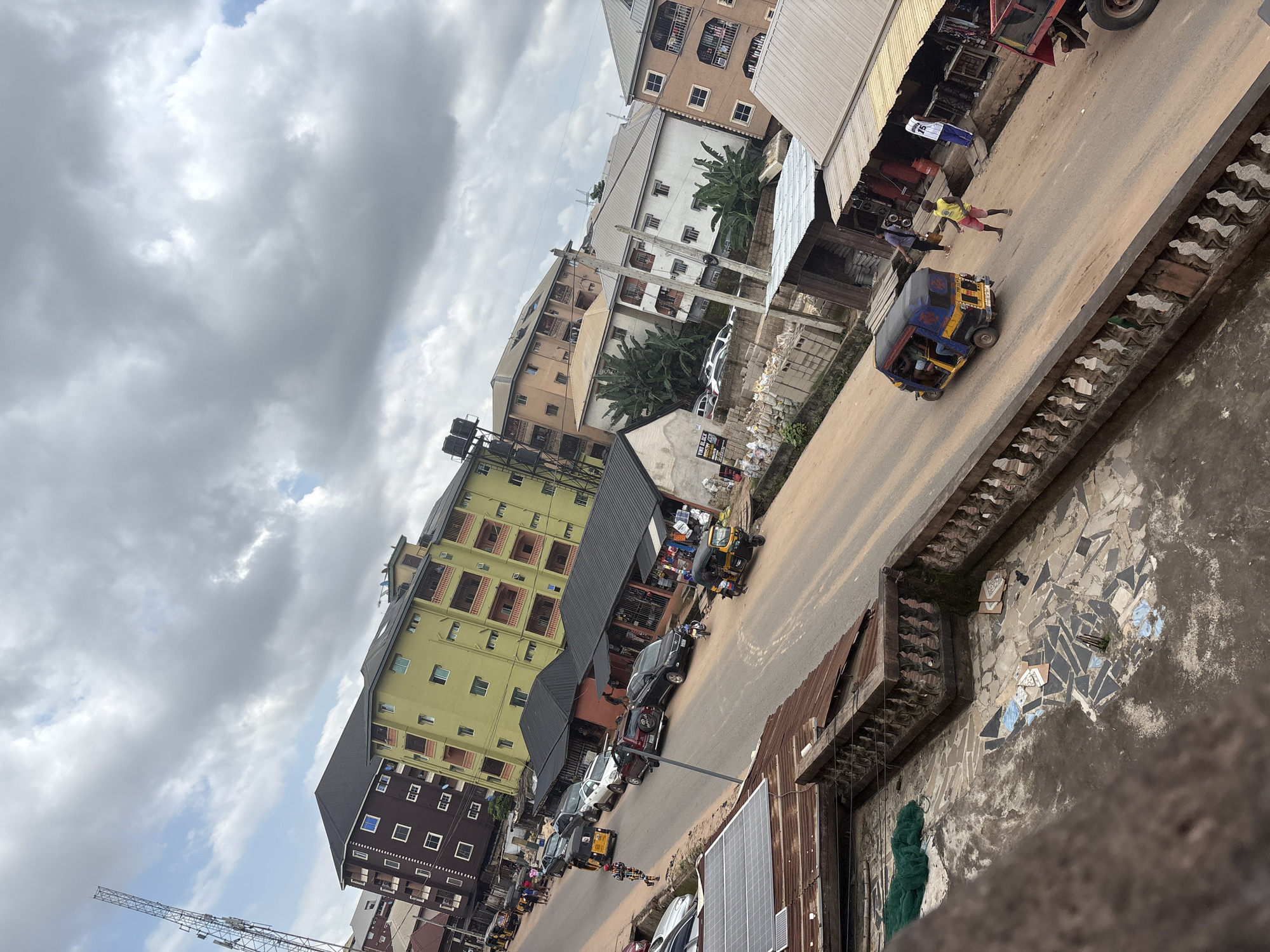 The image depicts a street scene in Nkpor, Nigeria, characterized by a two-lane asphalt road lined with various buildings and vehicles under a cloudy sky. On the left side of the road, a tall telecommunications tower stands prominently against the sky, followed by multi-story buildings with dark brown and lime green facades. Several cars are parked along the roadside, including a white sedan and a dark SUV. A yellow and blue auto-rickshaw is seen moving away from the camera.
Further down the road, a row of single-story shops with dark, sloped roofs are visible, displaying goods outside. One shop has a red gas cylinder next to it. A yellow auto-rickshaw is approaching from the right, with a driver and at least one passenger visible. Two individuals are standing near the shops; one in a yellow shirt and red shorts, and another wearing a white shirt with the number "15" on the back and blue pants. A red truck is partially visible on the far right of the frame.
On the right side of the road, there are more buildings of varying heights and colors, including a tan building with balconies and a white building with banana trees planted in front. A smaller, makeshift structure with a metal roof is situated closer to the road. The foreground shows a weathered balcony with ornate railings, a section of a tiled floor, and a green net. The overall scene suggests a busy, commercial area within a Nigerian city.