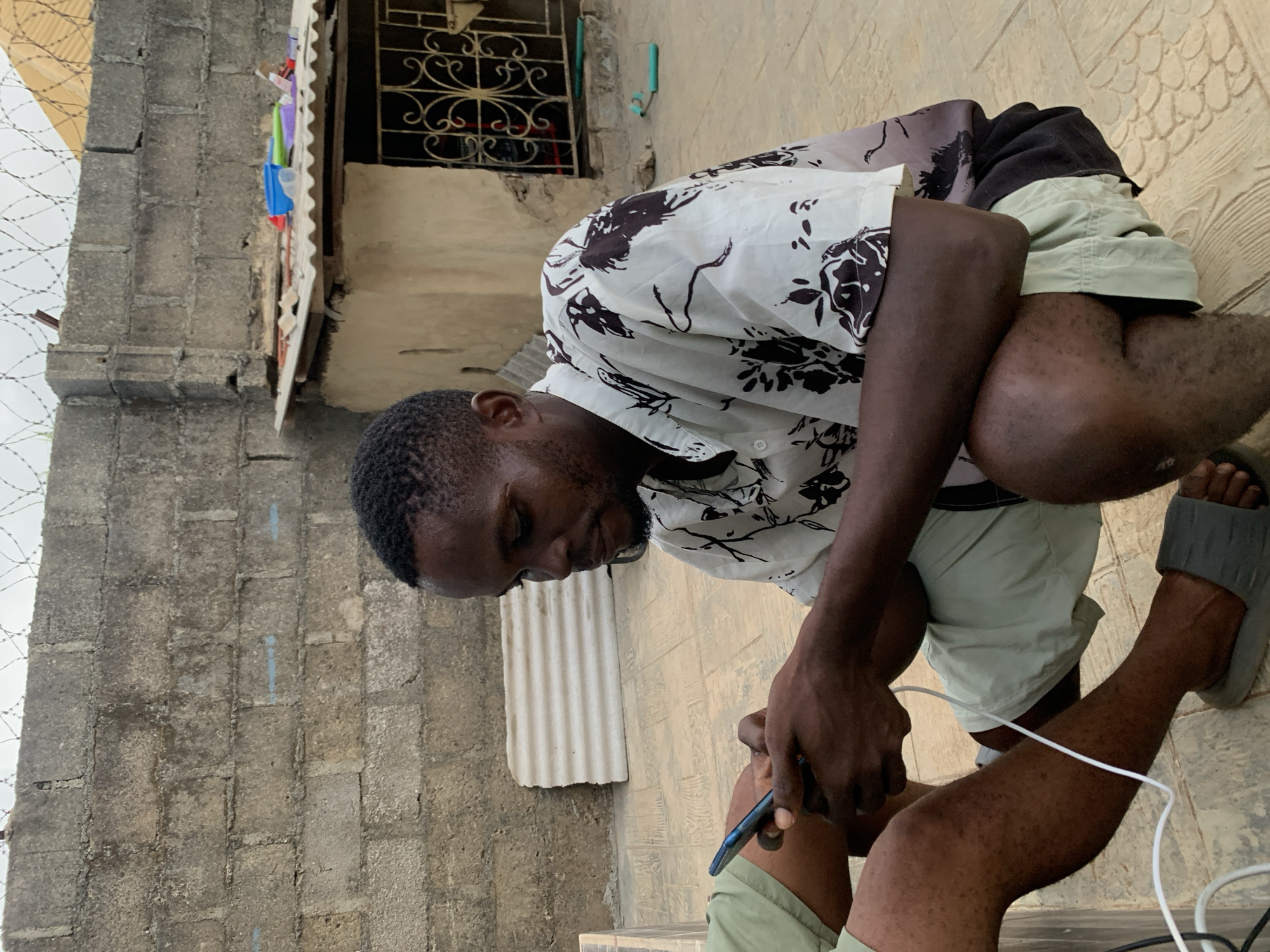 An African male squats on a textured surface next to a block wall in Ikorodu, Nigeria. He is wearing a white, short-sleeved shirt with dark brown floral patterns, light green shorts, and gray sandals. He holds a dark-colored smartphone. A white charging cable runs from the phone. The background shows a building with a decorative metal window covering and barbed wire along the top of the wall.