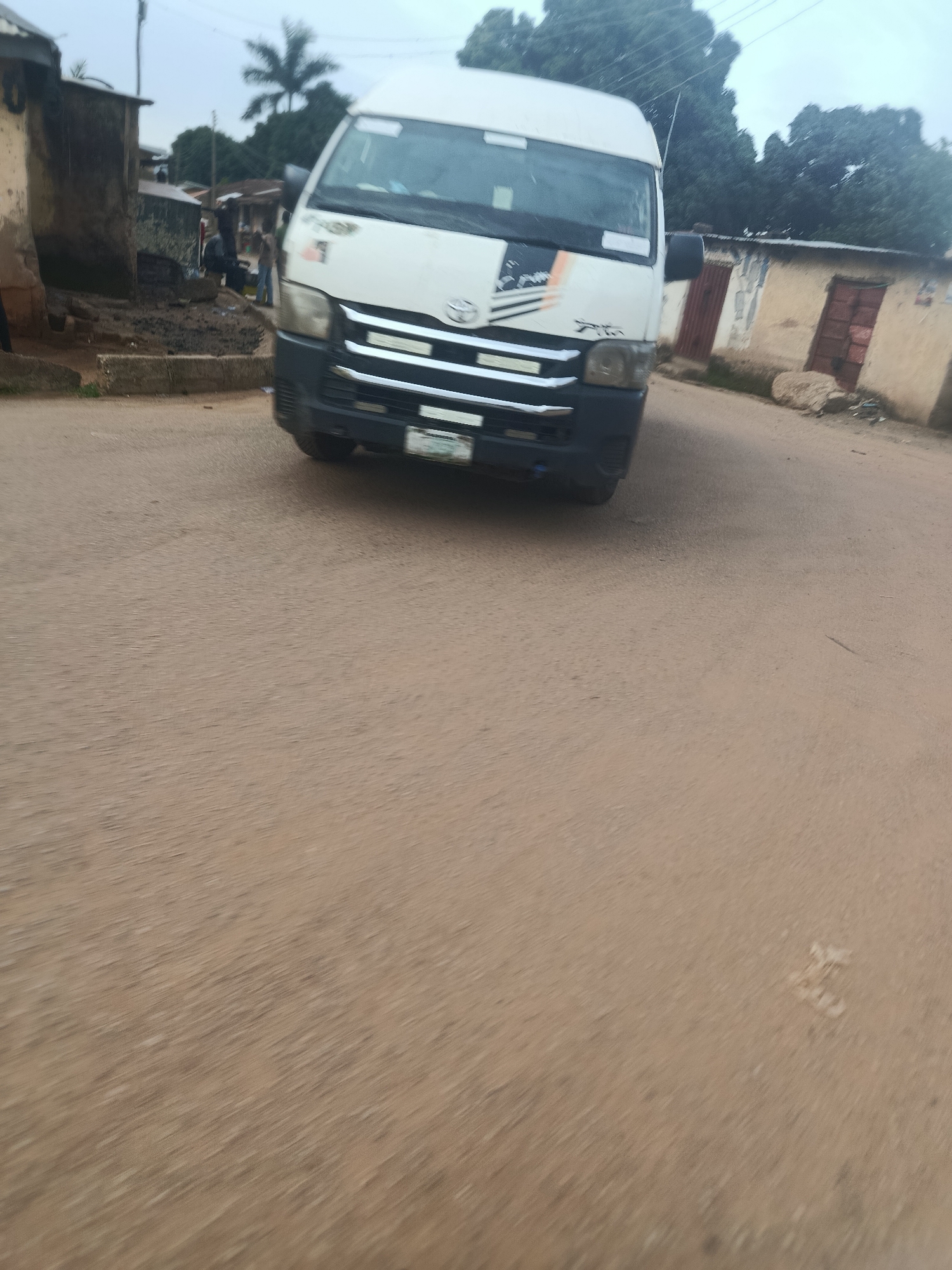 A white minibus is parked on a dusty road in Rafin Par, Nigeria. The minibus has a black and orange stripe on its side and a Toyota logo on the front. Buildings with corrugated iron roofs are visible on either side of the road. A palm tree stands tall in the background. The road is unpaved and appears to be in a rural area. The sky is overcast, suggesting it might be a cloudy day.