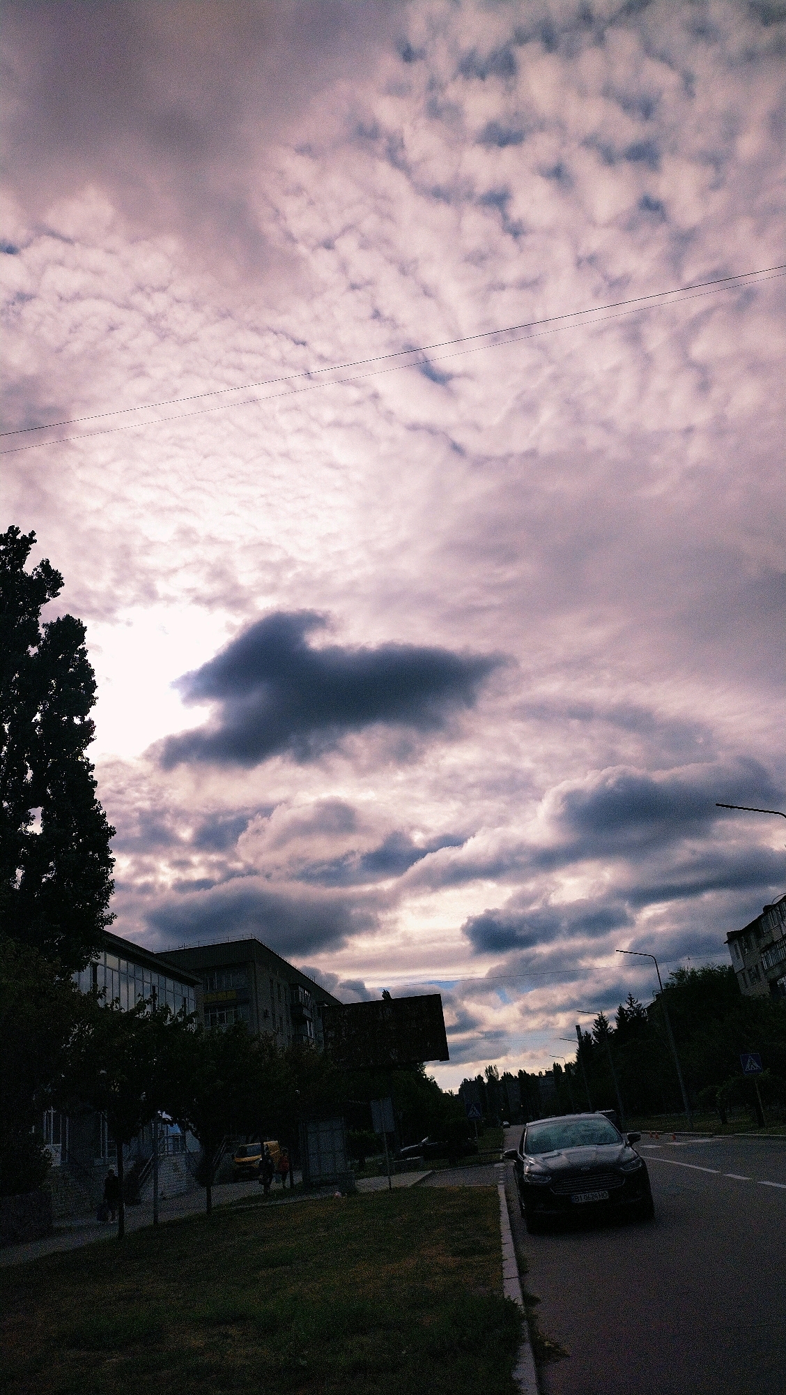 The image depicts an overcast sky with scattered clouds, predominantly a mix of light and dark gray with hints of pinkish hues, suggesting either sunrise or sunset. Below the sky, a treelined street runs parallel to a sidewalk. On the sidewalk, several people are visible walking. To the left of the street, buildings are present, one appearing to be a multi-story apartment building. A dark-colored car is parked on the side of the road. The overall atmosphere is muted, likely due to the cloudy weather. The scene is consistent with an urban setting in Horishni Plavni, Ukraine, with its typical residential buildings and infrastructure.