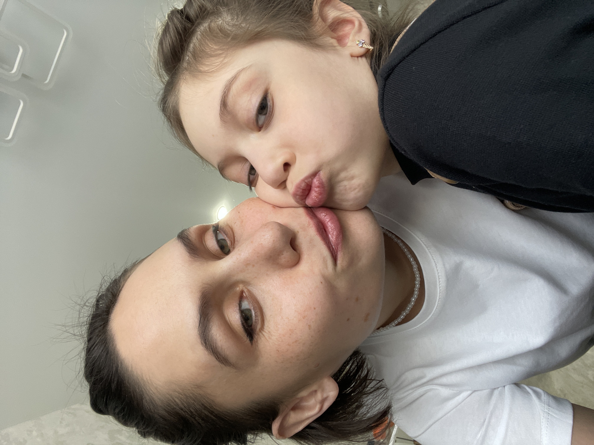 A close-up, slightly angled shot depicts a woman and a young girl in an affectionate pose. The woman, with short dark hair, fair skin with freckles, and wearing a white t-shirt and a pearl necklace, is positioned at the bottom of the frame. The young girl, with light brown hair partially pulled back and wearing a black top, leans in to kiss the woman's cheek. The background is a plain, light-colored wall with indistinct lighting fixtures. No information on the location being in the city of Izhevsk, Russia, can be derived from this image.