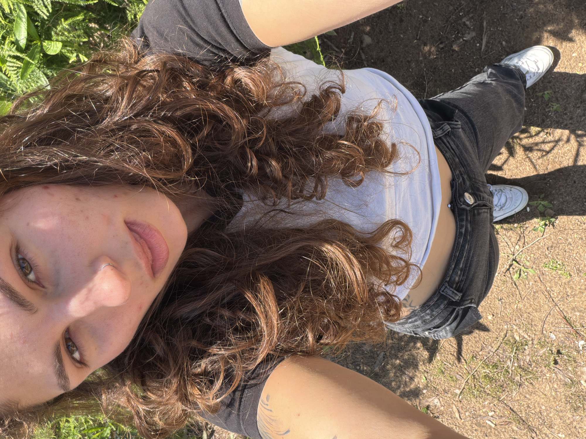 A young woman is taking a selfie outdoors. She has curly brown hair and is wearing a white t-shirt layered under a black top and black jeans. She is standing on an area of dry ground covered in sparse vegetation. Her white shoes are visible on the ground. The background is blurred, with glimpses of greenery suggesting a park or garden-like setting. The angle suggests the photo was taken from a slightly elevated position, looking down at the subject. The presence of plants and the dry ground indicate an outdoor environment, which could be consistent with a location in Kiryat Bialik, Israel.