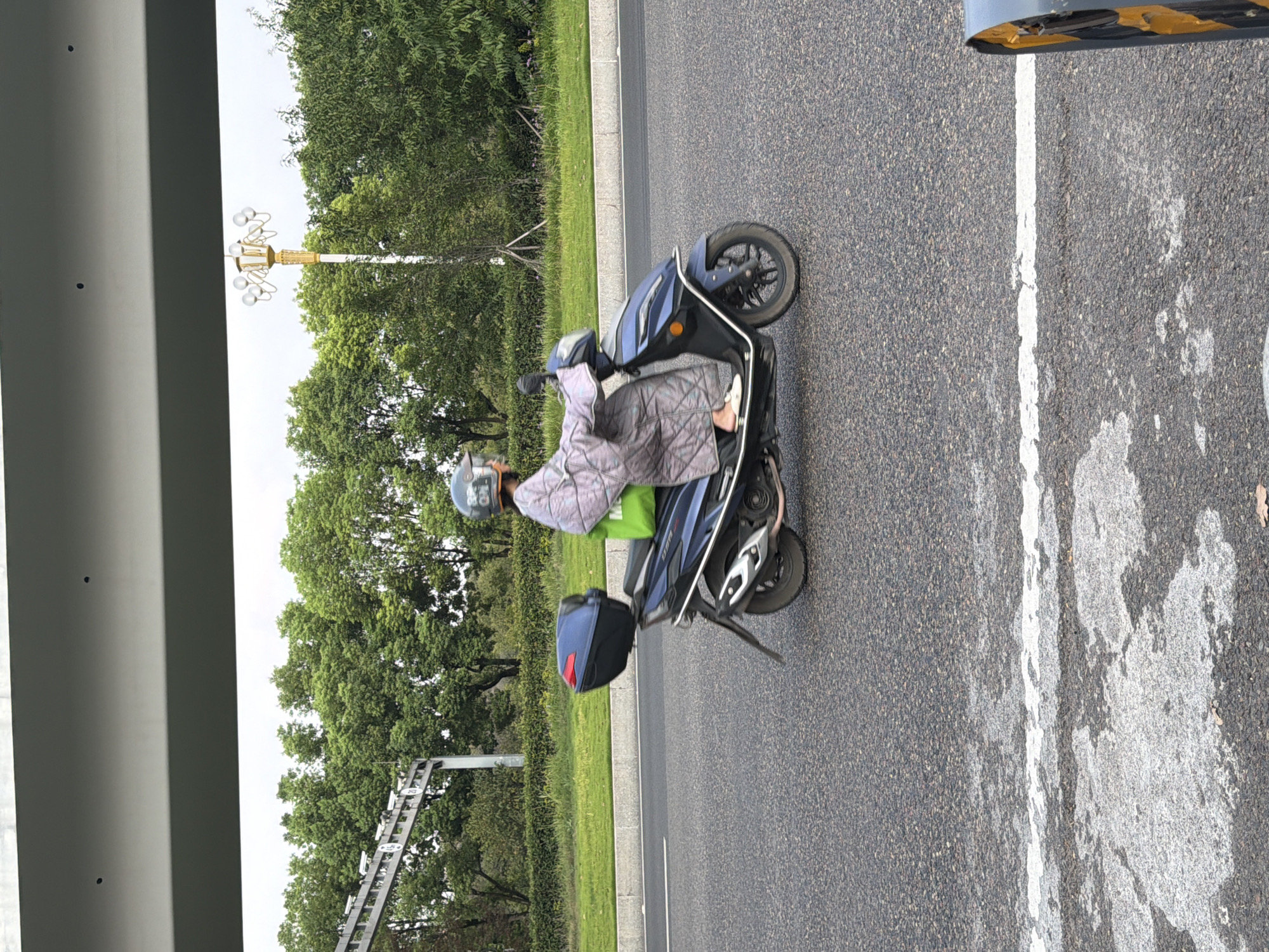 In Jiangjin, China, a person is riding a dark blue scooter on an asphalt road. The rider is wearing a light-colored quilted jacket with a patterned design and a gray helmet. A bright green bag is secured on the seat behind the rider, and a dark blue storage box is mounted on the back of the scooter. The scooter has a silver exhaust pipe. The road is lined with lush green grass and trees. Overhead, a portion of a white structure, possibly a bridge or overpass, is visible. A street lamp with multiple bulbs stands among the trees. The sky is overcast. The road surface shows markings and some water stains.