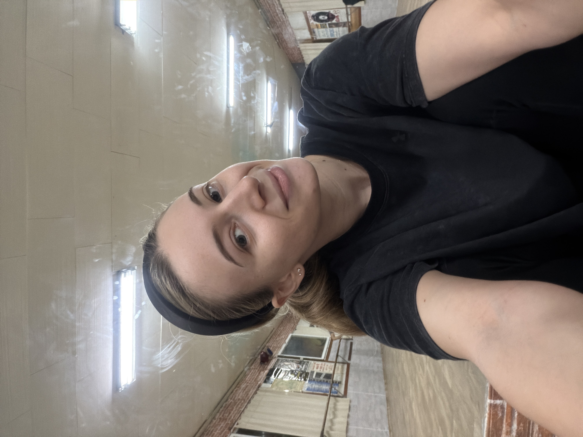 A woman with light skin, dark eyebrows, and light brown hair pulled back by a black headband is facing the camera, smiling slightly. She is wearing a dark-colored T-shirt. The background reveals a tiled hallway with fluorescent lights on the left. Windows and brickwork are visible on the right side of the hallway. Based on the architecture and design elements, the location appears to be in Plovdiv, Bulgaria.