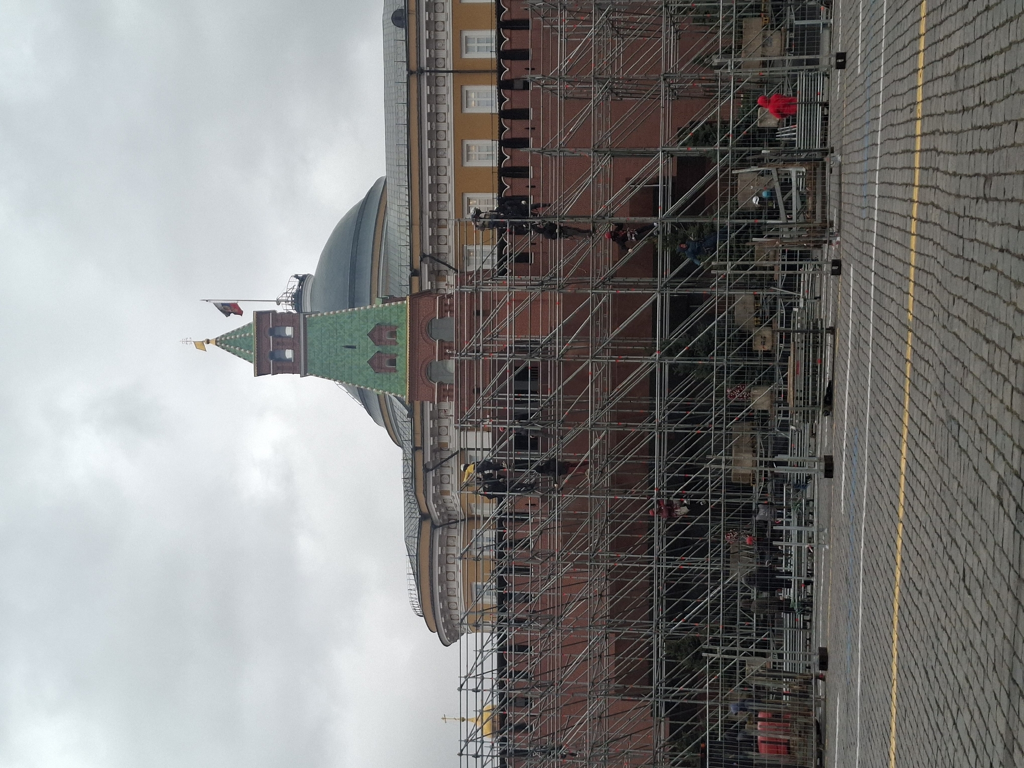The image displays a large historic building, identifiable as part of the Moscow Kremlin complex, partially obscured by extensive scaffolding. On the left, a prominent brick tower with a green-tiled, conical roof is visible. A flagpole on this tower flies what appears to be the Russian flag (white, blue, red horizontal stripes). Behind the tower, a large dome and a building with yellow upper walls and white-framed windows can be seen.
The building's facade is covered by a substantial structure of silver metal scaffolding, suggesting ongoing construction, maintenance, or setup for an event. Several individuals, likely workers, are positioned on different levels of the scaffolding. One person stands out in a bright red jacket, while others are visible wearing hard hats, including blue and yellow ones.
To the right, a broad expanse of grey cobblestone pavement is visible, marked with parallel yellow and white painted lines. This surface is characteristic of a major public square, hinting at preparations for a parade or large-scale gathering. The sky above is uniformly overcast and grey, indicating a daytime scene under cloudy weather conditions. No discernible text is present in the image.