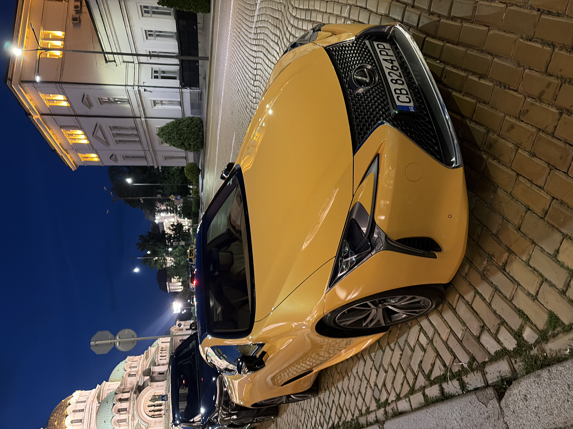 The image depicts a night scene in Sofia, Bulgaria, centered on a bright yellow Lexus LC 500 convertible parked on a cobblestone street. The car's front is angled towards the viewer, displaying its distinctive spindle grille, Lexus emblem, and a Bulgarian license plate reading "CB 8264 PP" framed with "Lexus" branding. The vehicle appears clean and reflective, illuminated by streetlights and ambient city light.
Behind the Lexus, a dark-colored SUV is partially visible, also parked. In the far background to the left, the prominent gold and green domes of the St. Alexander Nevsky Cathedral are visible, characteristic of its Byzantine Revival architecture. To the right of the image, a large, multi-story white building with ornate details and brightly illuminated windows stands, reflecting warm yellow light.
The street itself is paved with light-colored cobblestones, with some grass growing between the stones near the curb on the left side. Streetlights are visible throughout the scene, casting light on the buildings and the roadway. The sky is dark blue, indicating either late dusk or nighttime. No individuals are explicitly visible in the frame. A yellow taxi can be seen in the distance, further down the street to the right. The overall atmosphere is urban and well-lit at night.
