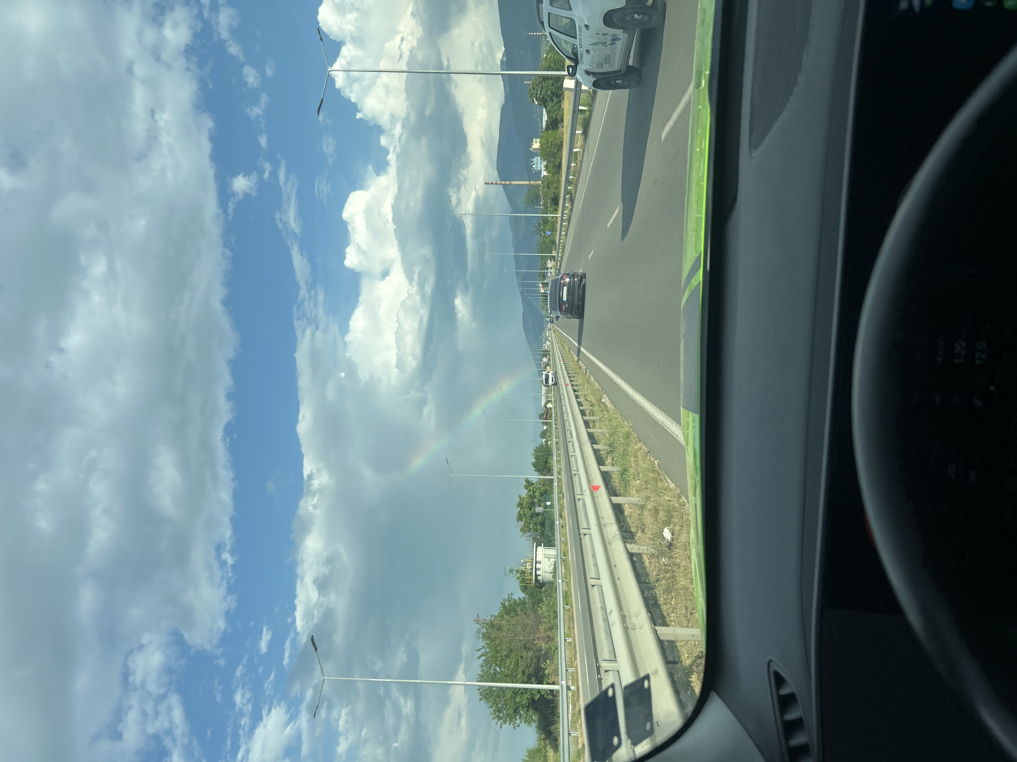 The image captures a view from inside a vehicle traveling on a multi-lane highway under a partly cloudy sky. The perspective is from the front passenger side, with the edge of a steering wheel and dashboard visible on the right.
The sky features a mix of bright blue patches and large, fluffy white clouds, with darker, heavier clouds towards the left horizon. A faint rainbow arcs across the sky on the left side of the highway, suggesting recent rain or misty conditions in the distance.
The highway stretches into the distance, lined with guardrails and utility poles. On the right-most lane in the foreground, part of a white SUV or truck is visible. Further down the road, a dark car is in the center lane, and other vehicles can be seen in the distance. The highway appears to have a distinct green-colored lane or shoulder on its immediate right side.
In the background, mountains rise under the cloudy sky, and various structures, including a tall, slender chimney or smokestack and other buildings, are visible among trees.
The time of day appears to be daytime, with ample sunlight. The weather is mixed, characterized by both clear skies and clouds, along with the rainbow. On the visible part of the dashboard/steering wheel on the right, numbers such as "100" and "120" are faintly legible, likely part of a speedometer or tachometer.