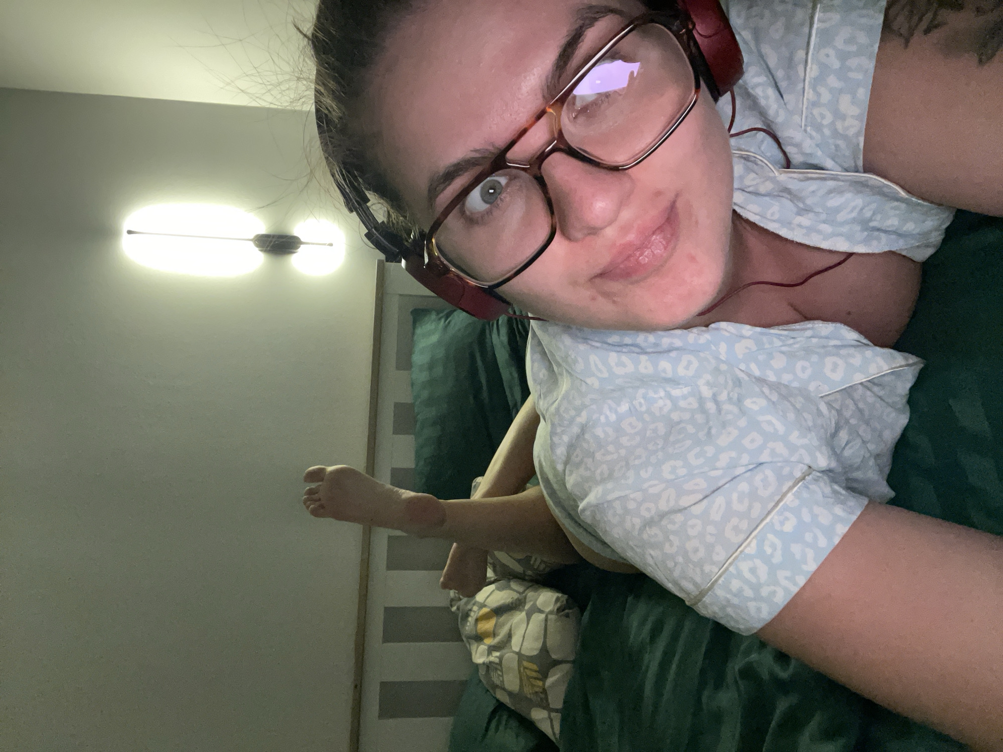 A woman lies on a bed, wearing glasses and headphones. The bed has a green comforter and pillows. One of her bare feet is visible in the background. A light fixture is on the wall behind her. The location is not discernible from the image, so a location in Мосметаллоконструкция Онт, Russia cannot be confirmed.