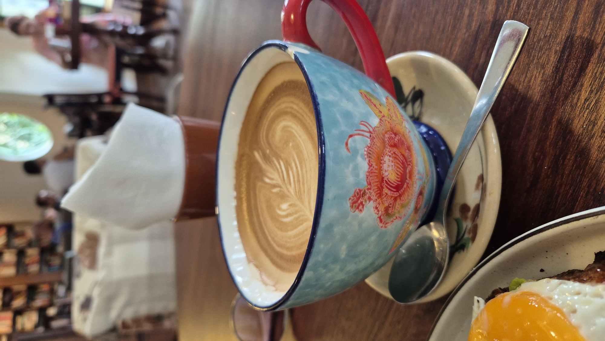 A close-up shot captures a beautifully decorated coffee cup filled with latte art on a wooden table. The cup, adorned with a vibrant floral pattern, features a red handle. A spoon rests on the saucer beside it. In the background, out of focus, are bookshelves lining a wall, suggesting a cafe or library setting. The lighting is soft, casting gentle shadows. The scene appears to be set in a cafe in Da Nang, Vietnam, hinted at by the overall ambiance and the possibility of a local establishment.