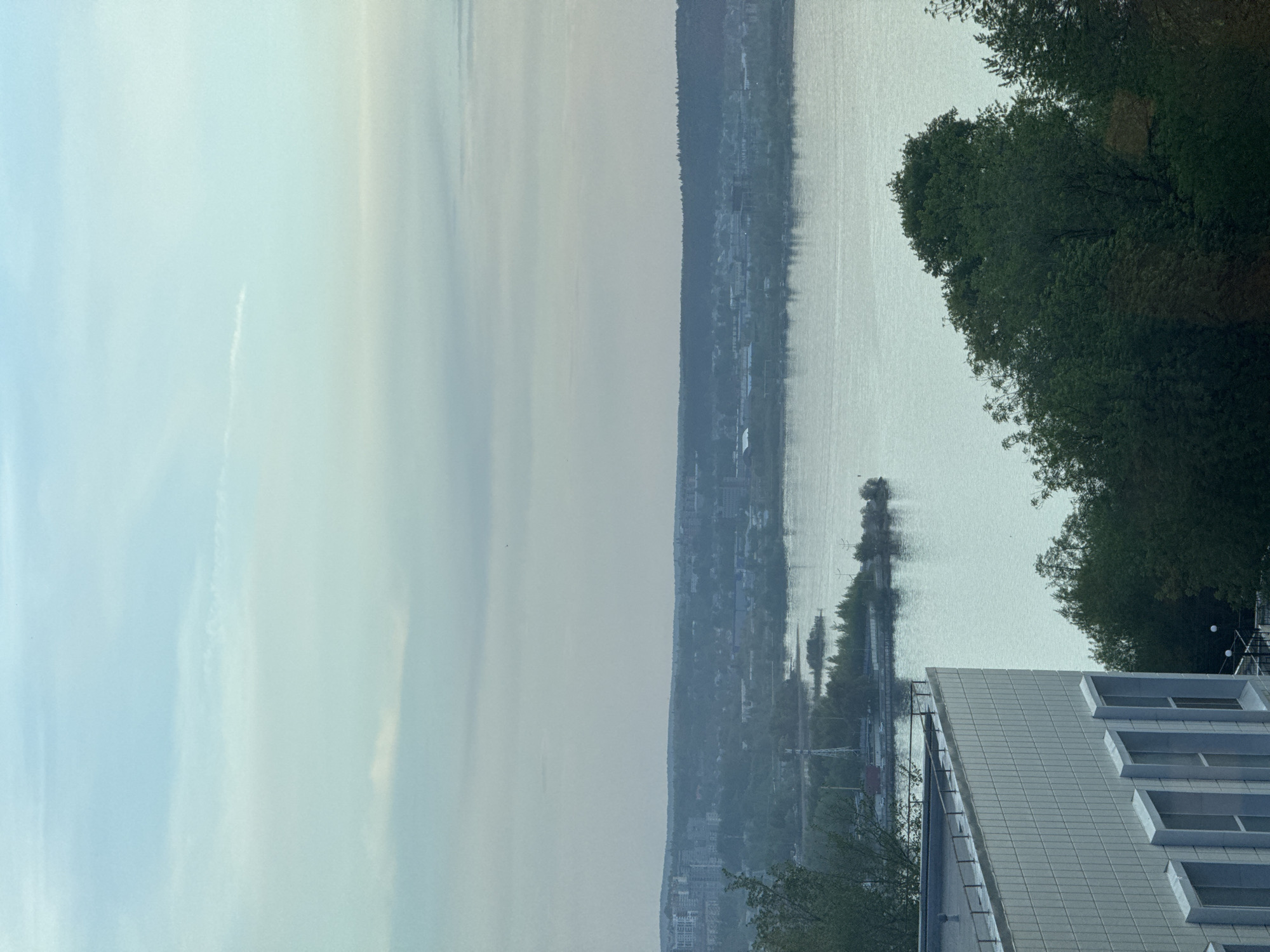 A long body of water is visible in this image, likely a river or lake, bordered by a shoreline with trees and buildings. In the distance, a town or city is situated on the opposite shore. The sky above is overcast, and the light is diffused, suggesting it might be early morning or late evening. In the foreground, a building with multiple windows is seen; a tree is visible as well. Given the description, this might be located in Izhevsk, Russia.