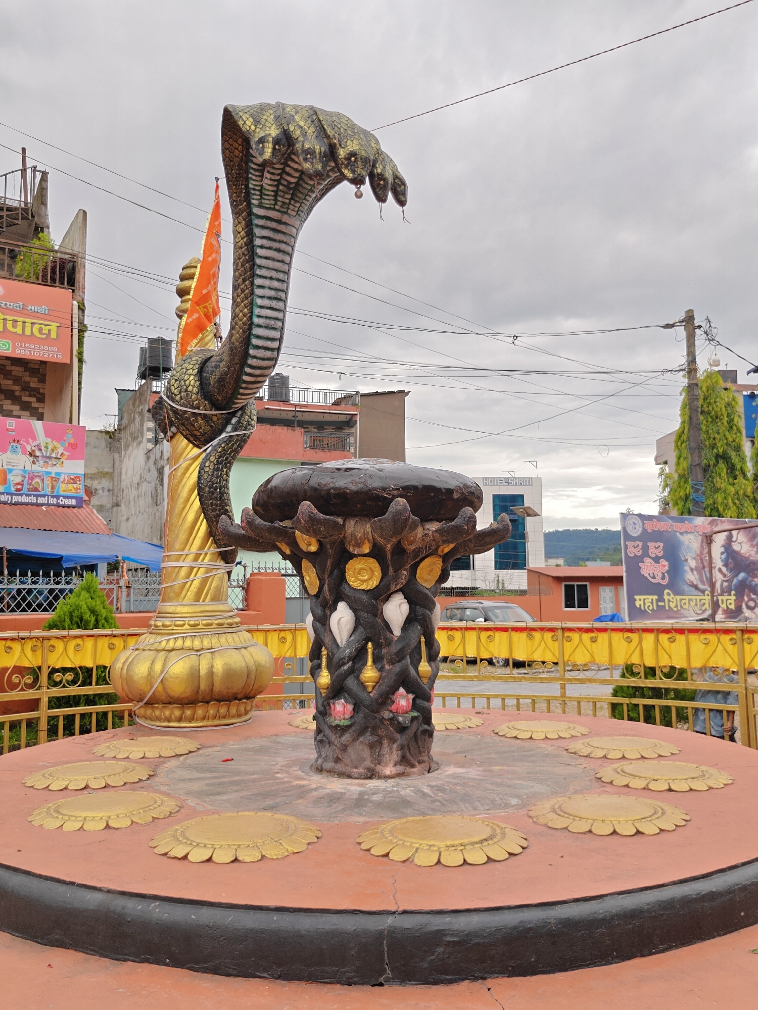 The image shows a statue of a multi-headed cobra in Pokhara, Nepal, likely a religious or decorative monument. The cobra statue is a prominent feature in the foreground, with its scales detailed and its hoods spread. It is positioned atop a dark, textured base decorated with white shell-like objects and possibly lotus flowers. A golden pillar, partially obscured by the cobra's body, supports another element of the statue or a surrounding structure. An orange flag with writing is visible behind the cobra. The statue is situated on a circular platform with a reddish-orange surface and decorative golden floral motifs. A yellow metal railing encloses the platform. In the background, buildings, trees, utility poles with wires, and a cloudy sky are visible, indicating an urban or semi-urban environment. A banner with religious imagery and text in what appears to be Nepali or Hindi is also present in the background. The overall scene suggests a public space in Pokhara, possibly a temple complex or a roadside attraction.