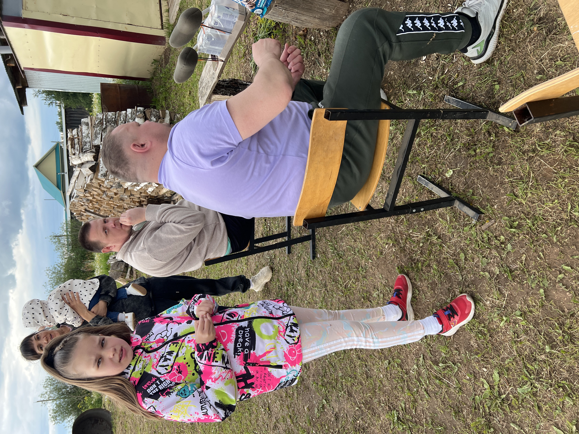 The image shows a group of people in what appears to be a rural or outdoor setting. A young girl is in the foreground, wearing a brightly colored jacket and red shoes. Behind her, several adults are seated, possibly on chairs or benches. One man is wearing a purple shirt and green pants with a distinctive snake-like pattern on the side. Wood and buildings are in the background, suggesting a countryside or village environment. Based on the location, this scene could be taking place in Большие Кусты, Russia.