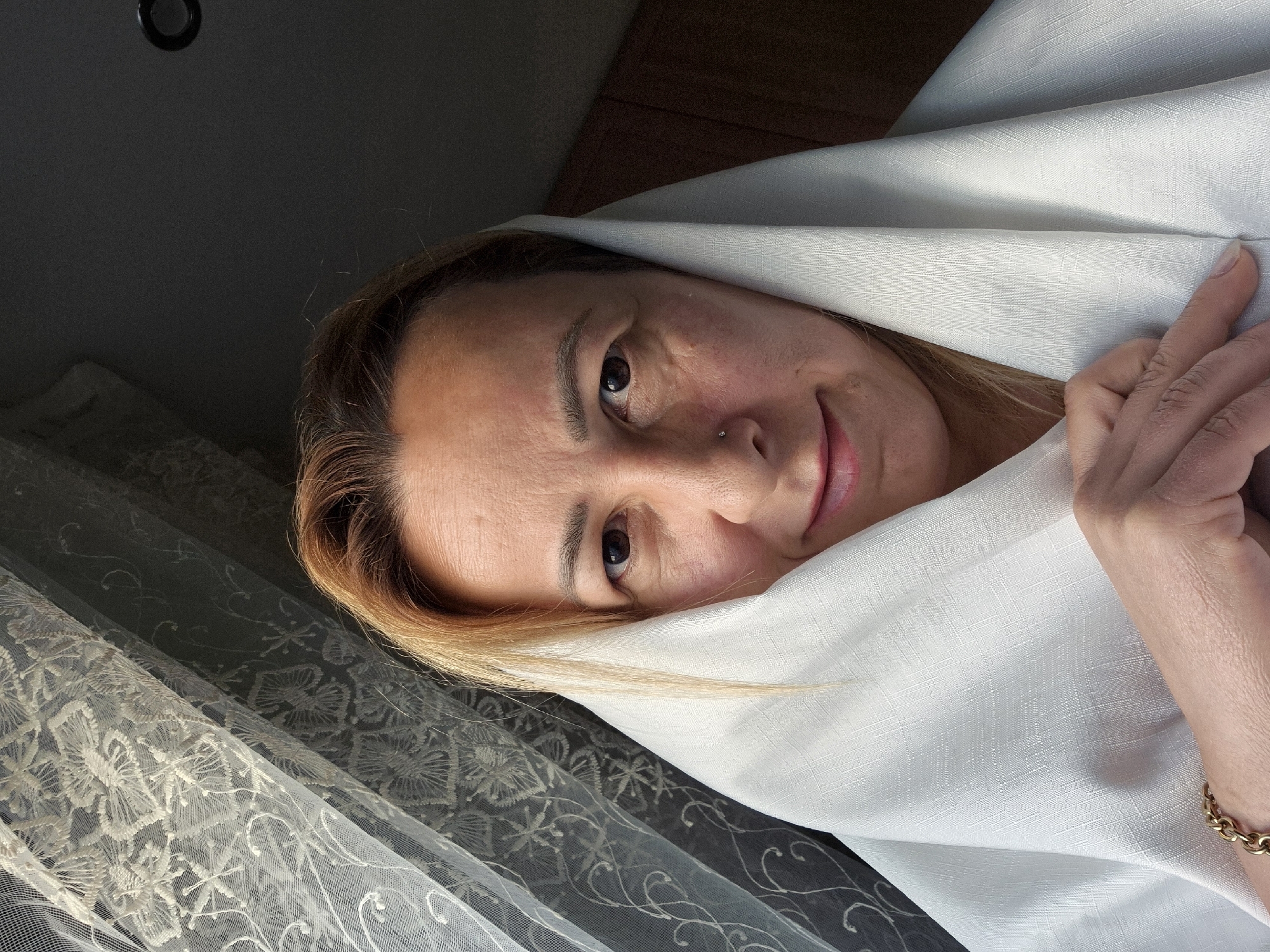 A medium shot portrait shows a woman with blonde hair wearing a white headscarf. She is looking directly at the camera with a slight smile. Her eyes are brown, and she has a small nose piercing. The background consists of a sheer lace curtain on the left and a dark wall with a wooden cabinet on the right. The lighting suggests it is daytime, with light coming from the left, possibly through a window. The woman's left hand is visible, resting on the headscarf, and she is wearing a gold bracelet. The scene appears to be indoors, in an unknown location.