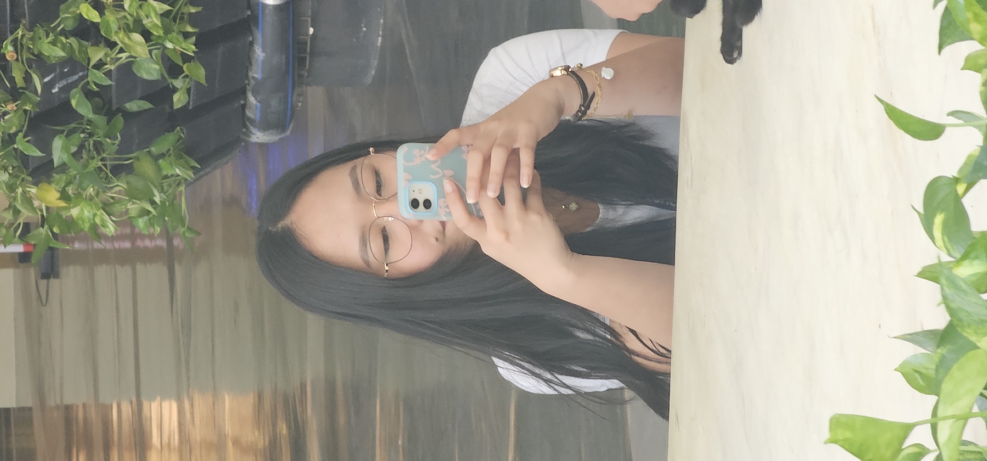 A young Asian woman with long black hair and glasses is taking a selfie in a mirrored surface, possibly a window or a polished wall. She is holding a light blue phone with a floral pattern. She is wearing a white t-shirt and several bracelets on her left wrist. Behind her, there is a large planter with green leafy plants. The background appears to be an indoor space with a polished, reflective floor. The presence of plants and the reflective surface suggest an indoor setting in a commercial or public space within Makati, Philippines, potentially a mall, hotel lobby, or office building atrium.