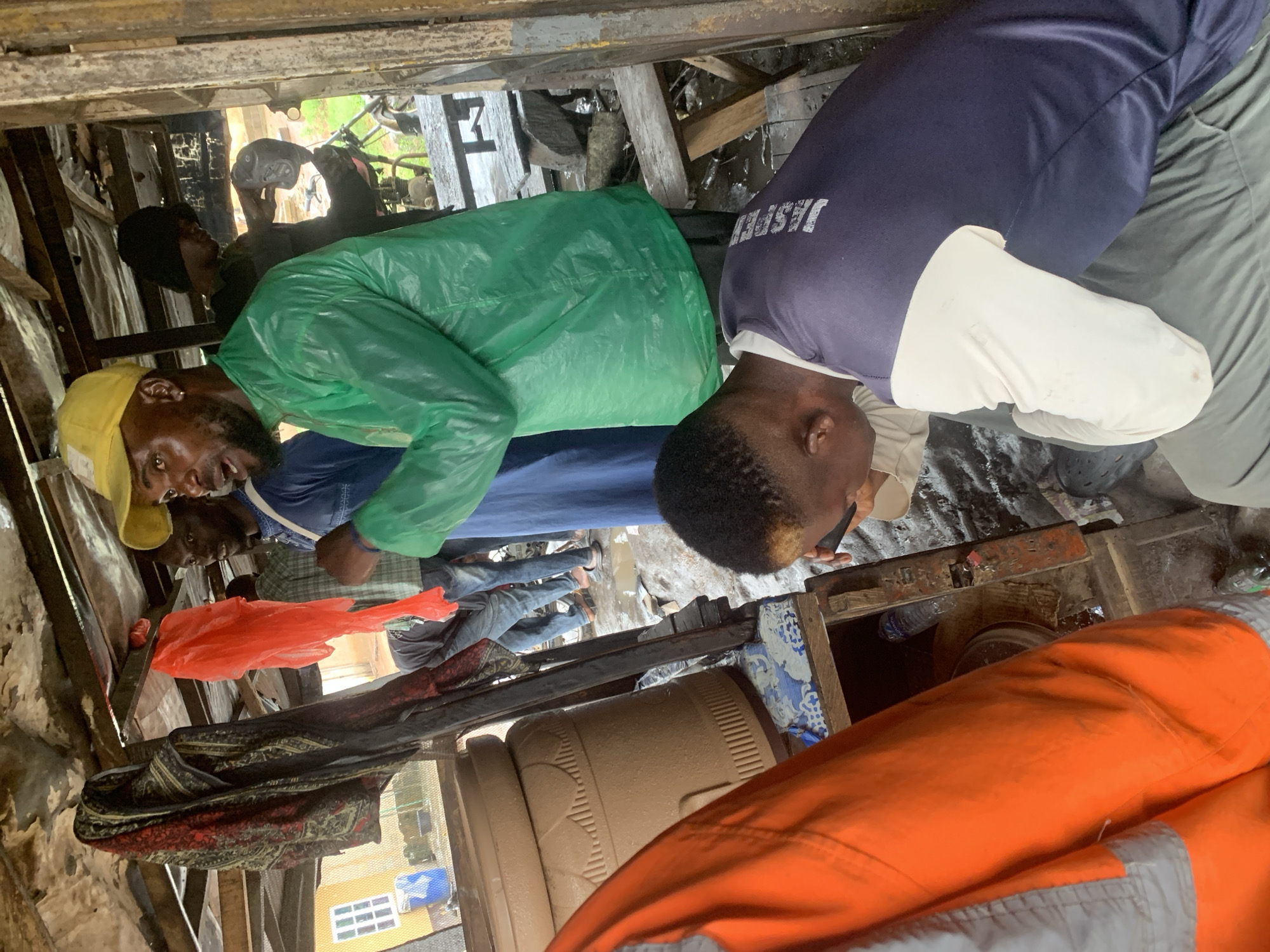 In Shagamu, Nigeria, a group of individuals are gathered within a makeshift structure. The scene appears to be either a market stall or a repair shop, with various items scattered around. The roof of the structure is made of corrugated metal and appears to be in disrepair.
In the foreground, a person wearing an orange and white jacket is hunched over, seemingly examining something on a wooden surface. To their right, another person in a green rain jacket and a yellow cap is looking towards the left. Behind them, several other individuals are visible, some wearing casual clothing, others in what might be work attire. One person is wearing a blue shirt and jeans, while another is wearing a black head covering.
Objects present include a large, beige water dispenser, a red plastic bag hanging from the ceiling, and several pieces of fabric. In the background, parts of a motorcycle and some wooden tables are discernible. The ground is littered with debris, suggesting an outdoor or semi-outdoor environment that may have experienced recent rain or a spill. The overall impression is one of a busy, possibly commercial, space in a developing urban area.