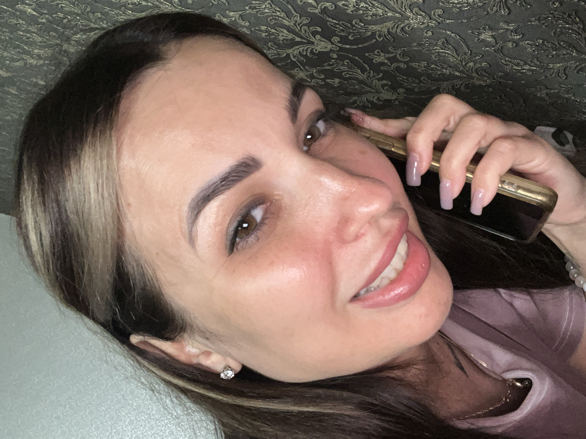 A close-up, high-angle, selfie-style shot shows a woman with brown hair and hazel eyes holding a gold-colored smartphone to her right ear while leaning her head against a dark, textured wall. The woman is wearing a pale pink shirt and a delicate gold necklace. Her lips are painted with a glossy pink lipstick, and she is smiling. She has long, light purple fingernails. The background wall has a repeating ornate pattern. The lighting is soft and casts a subtle glow on her face.