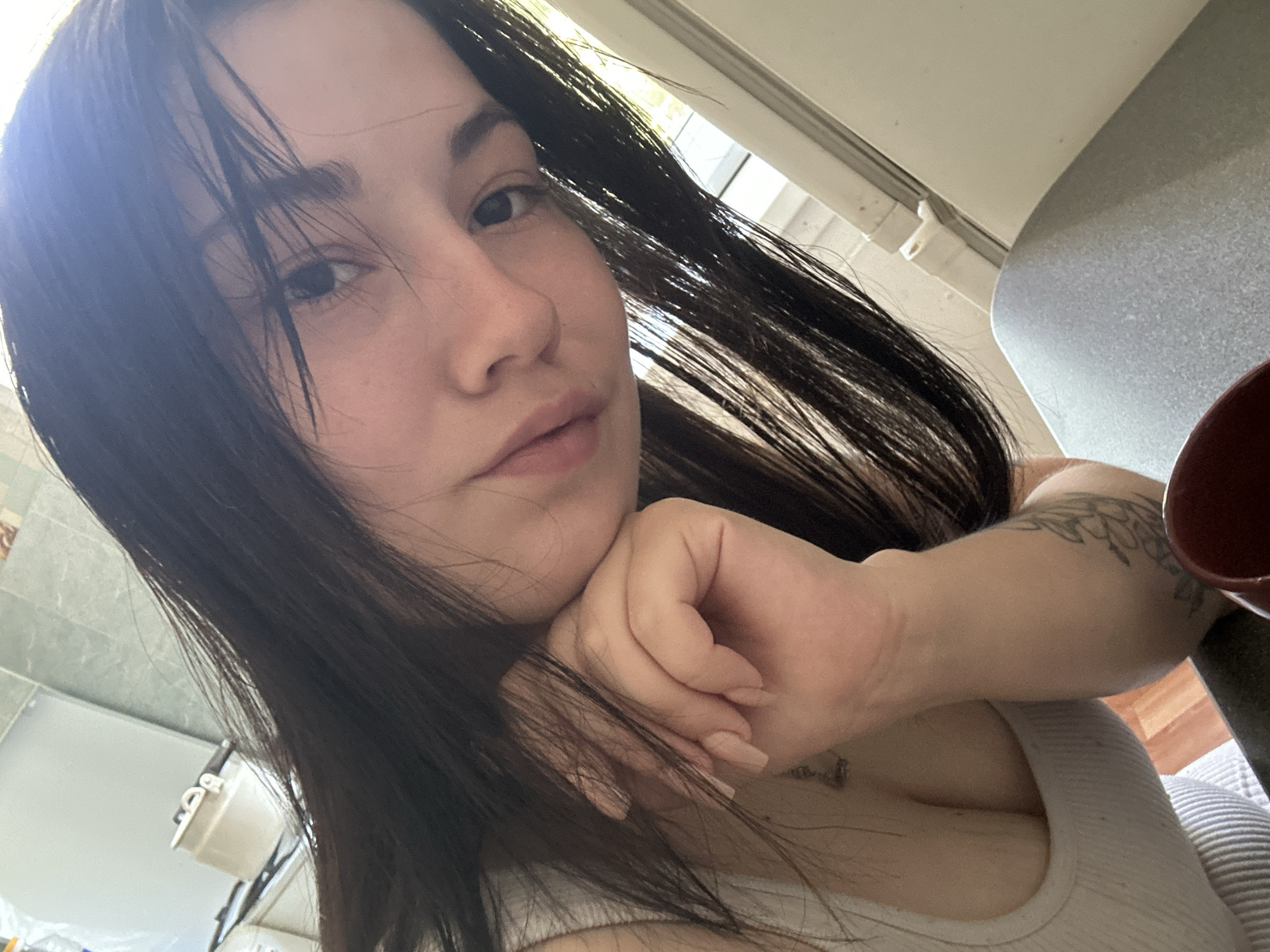 A young woman with long, dark hair is resting her head on her hand, looking towards the camera. She is wearing a white tank top, and her right arm is visible, adorned with tattoos. A portion of her chest is also visible. In the background, out of focus, are kitchen elements, including a tiled backsplash and what appears to be a kettle on a stovetop. The overall lighting suggests it is daytime. The image does not contain enough information to determine the specific location within Chaikovsky, Russia.