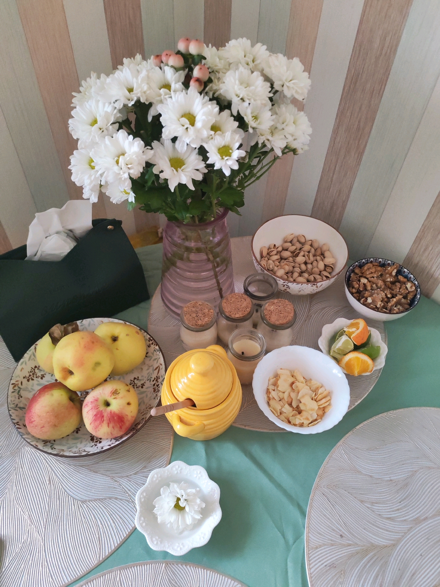 The image depicts a table set with various food items and decorative elements. A bouquet of white chrysanthemums in a pink glass vase is a prominent feature, placed on a surface with a striped, pastel-colored wallpaper in the background. To the left, a dark green tissue box is visible.
In front of the flowers, a patterned plate holds a few apples. Adjacent to the plate is a yellow ceramic honey pot with a wooden dipper. To the right of the honey pot, a small white bowl contains sliced almonds, and next to it, a white bowl holds citrus slices, including orange and lime. Further to the right, two white bowls contain nuts, one filled with pistachios and the other with walnuts. Three small glass jars with cork stoppers are also present, containing what appear to be candles.
In the foreground, a white floral-shaped dish holds a single white chrysanthemum. The table is covered with a light green cloth, and decorative placemats with a white, wavy pattern are placed on top.
The scene is a still life arrangement, likely indoors, with no indication of a specific city or location within Izhevsk, Russia. The image captures a moment of domestic arrangement, possibly for a meal or as a decorative display. There are no people or animals present in the image.