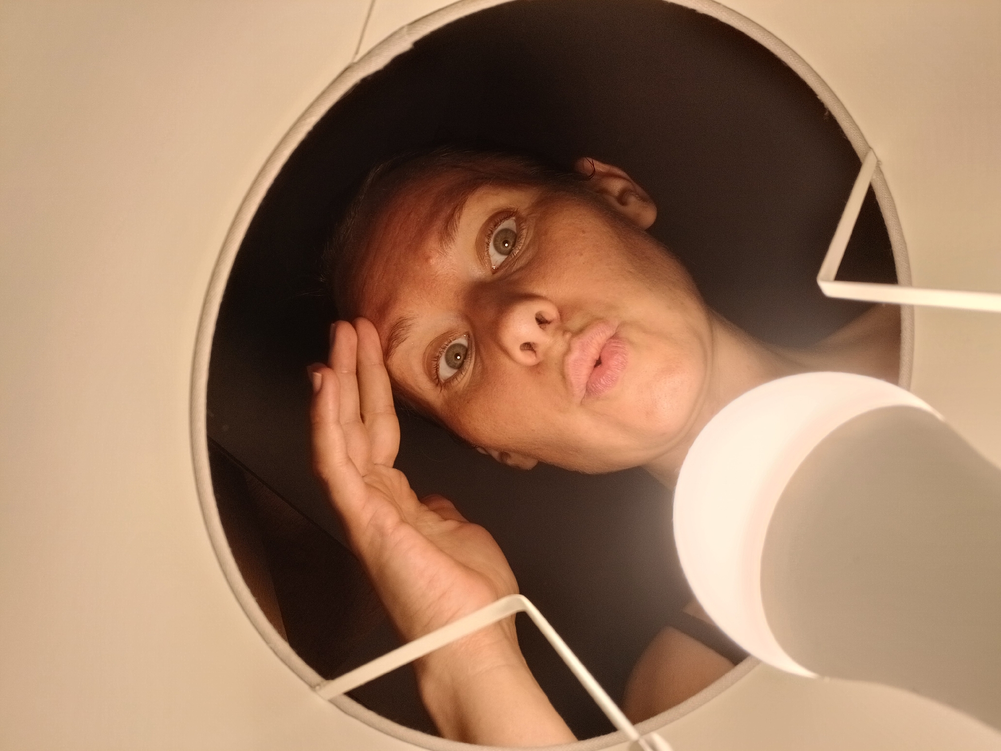 The image shows a person from a low angle, looking up into a lampshade. The person has their hand on their forehead and their mouth is puckered. A lightbulb is prominently featured in the foreground, partially obscuring the person's face. The background is dark and indistinct. The location is Yekaterinburg, Russia.